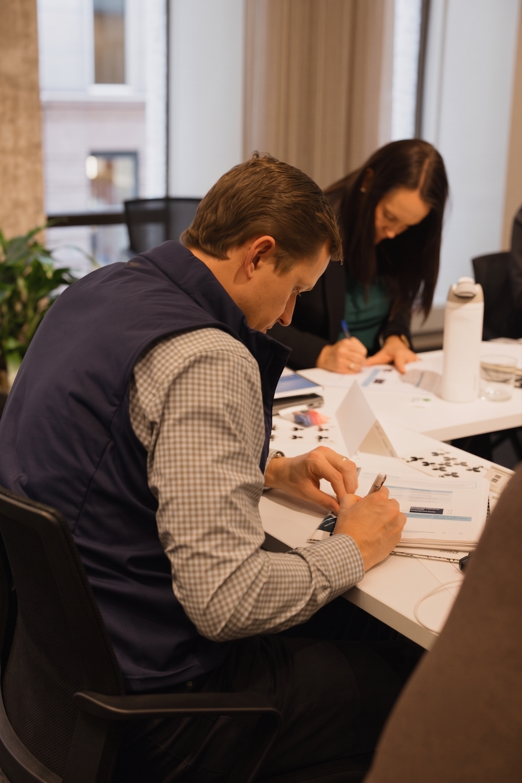 A man and a woman are working together at a table during a meeting or workshop, with papers, pens, and a water bottle.