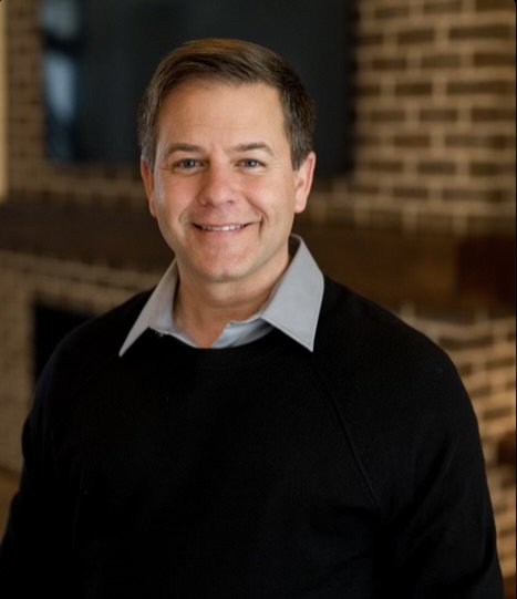 Portrait of a smiling man wearing a light blue dress shirt and black sweater, standing indoors with a brick wall in the background.