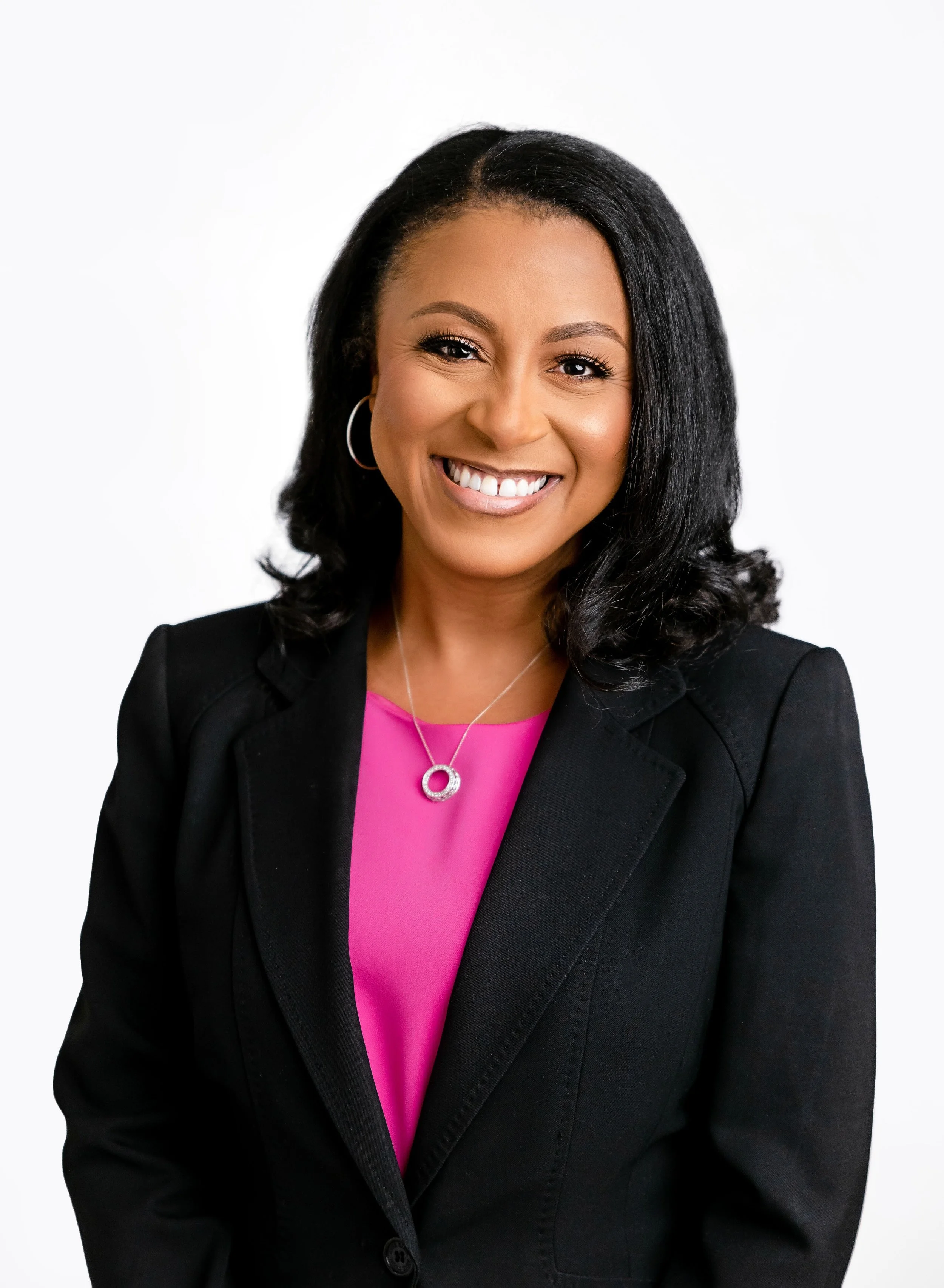 A smiling woman with curly black hair wearing a black blazer, pink blouse, and silver jewelry, posing against a white background.