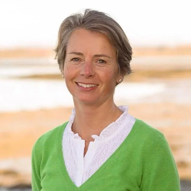 A woman with short gray hair smiling outdoors at the beach, wearing a white blouse and a bright green sweater.