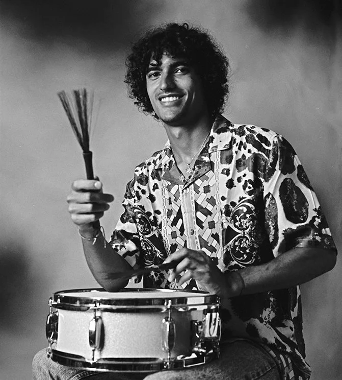Black and white photo of a young man with curly hair, smiling, holding drumsticks, seated behind a snare drum, wearing a patterned shirt.