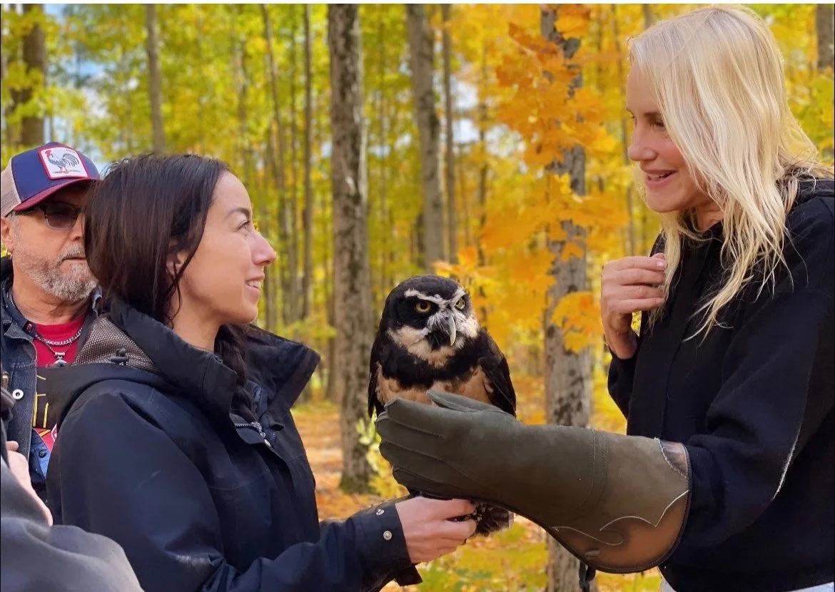 Two women are in a forest with autumn foliage, one is holding an owl and the other is smiling at it, while a man stands nearby observing.