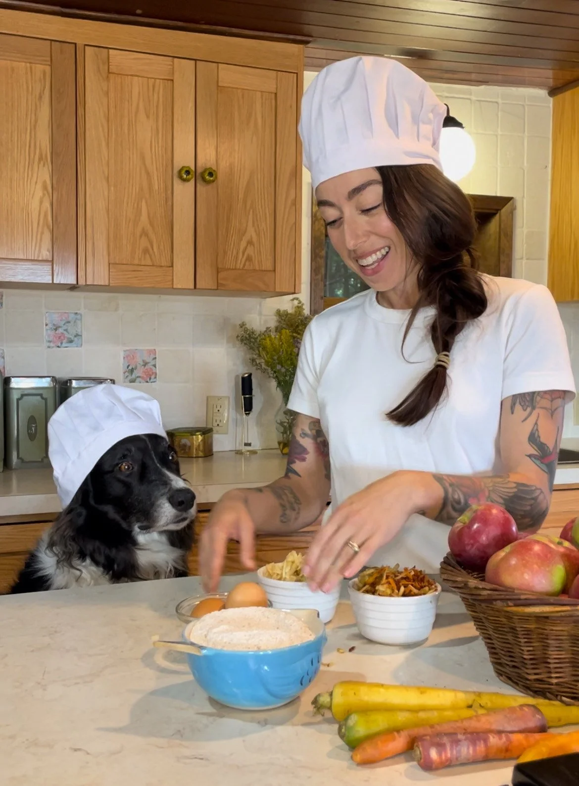 A woman and a dog wearing chef hats in a kitchen, preparing food on a countertop with apples, carrots, eggs, and bowls of ingredients.