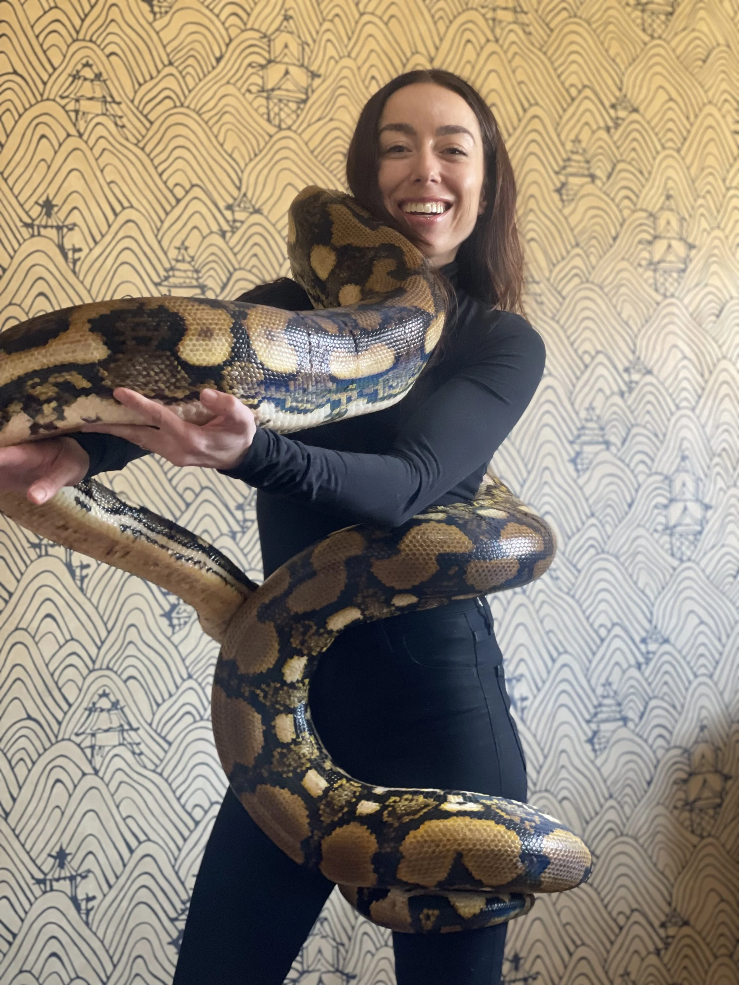 Woman smiling and holding a large snake in front of a patterned wall.