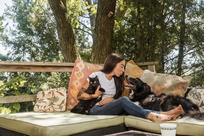 A woman sitting on an outdoor sofa with two dogs and a cat, surrounded by trees.