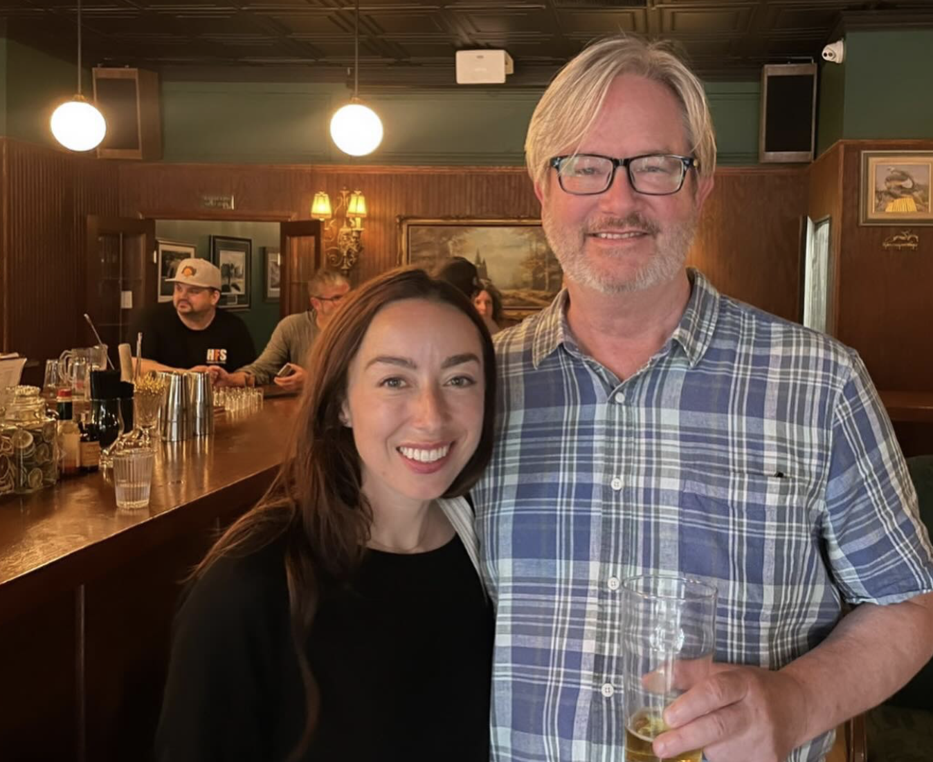 A smiling young woman with long dark hair and a middle-aged man with glasses and gray hair, holding a glass of beer, standing together in a cozy bar or pub with warm lighting, wooden decor, and framed pictures on the wall behind them.