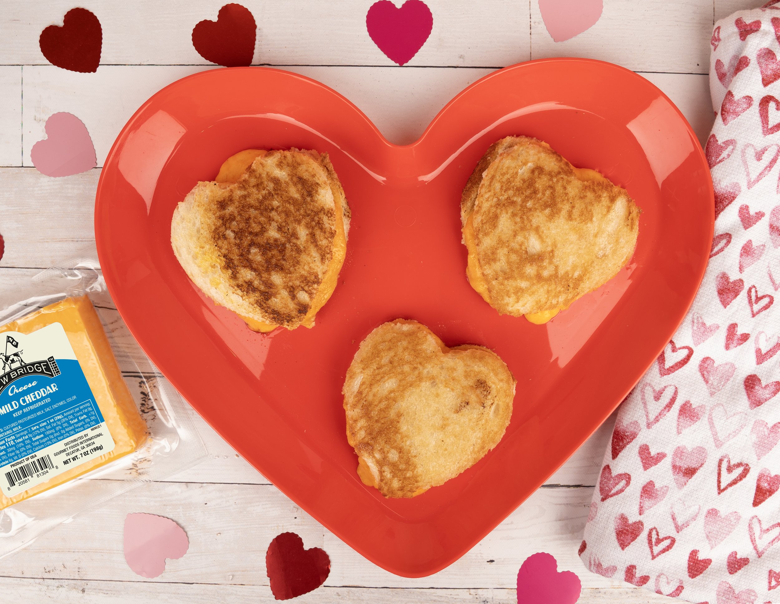 Three heart-shaped grilled cheese sandwiches on a red heart-shaped plate surrounded by pink and red heart-shaped confetti, with a package of New Bridge Mild Cheddar and a cloth with red and pink hearts on a whitewood surface.