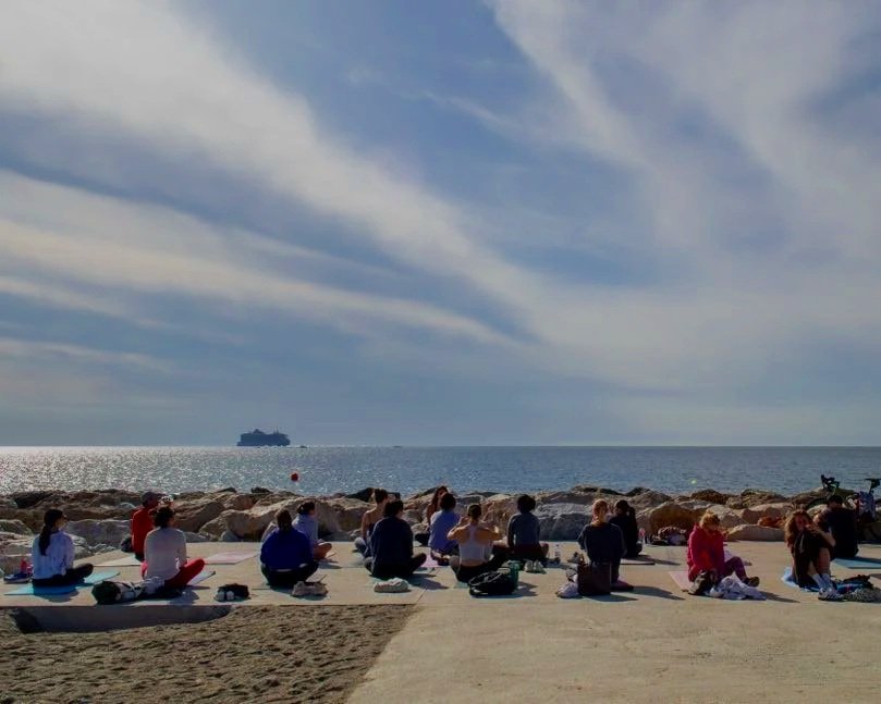 People practicing yoga on the beach during sunset, facing the ocean with a large ship in the distance.