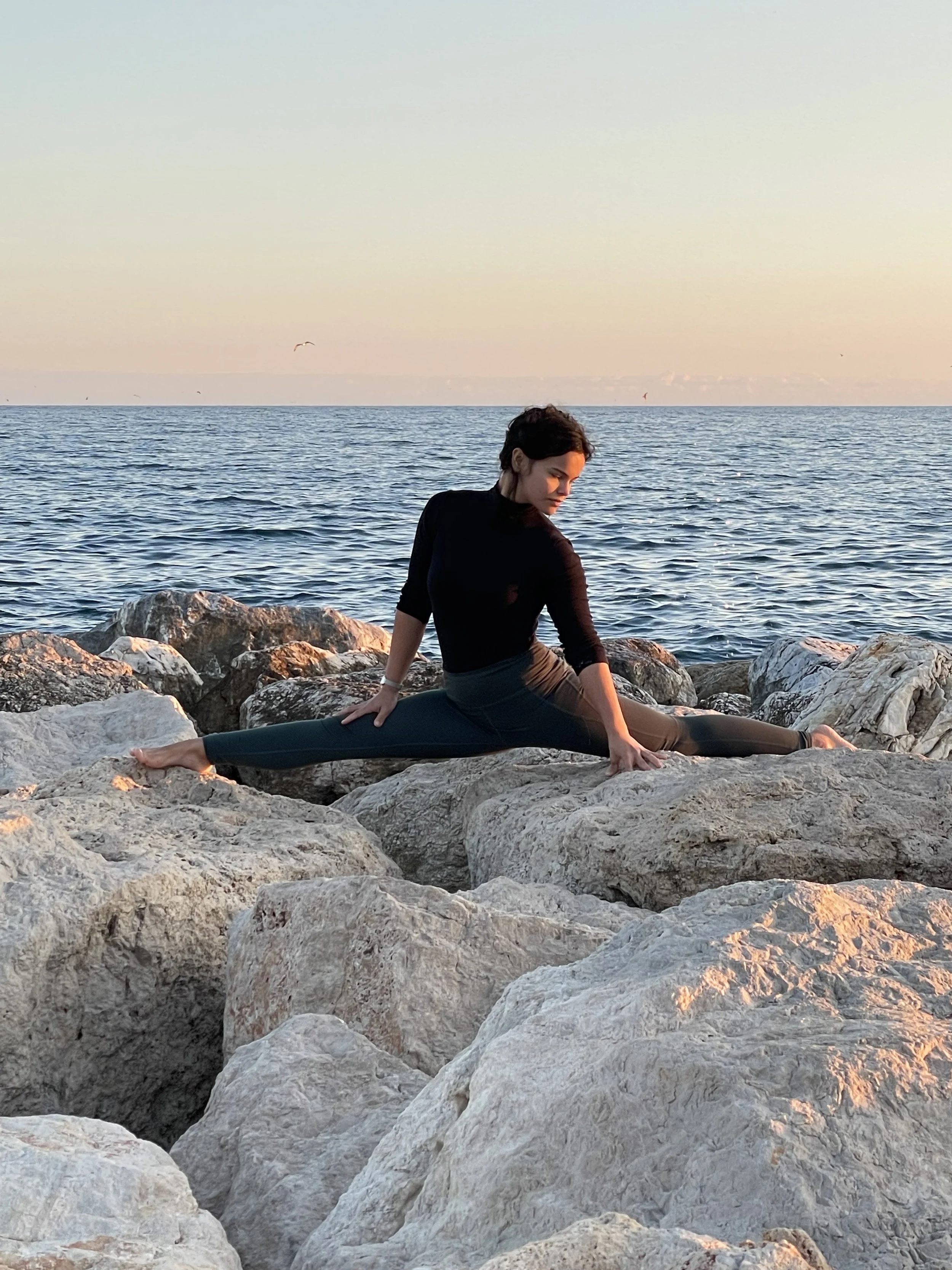 A woman performing a split stretch on rocks by the ocean during sunset.