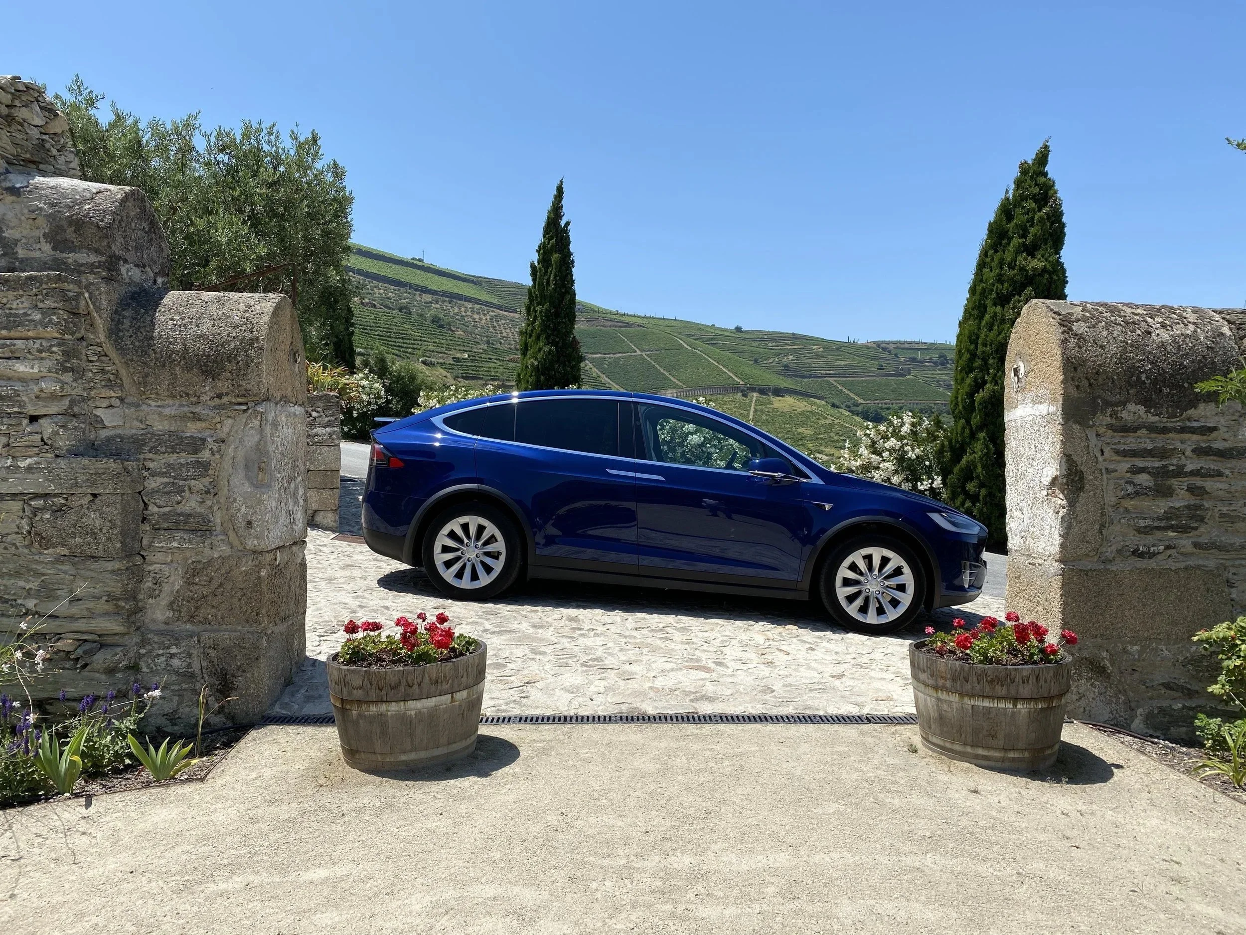 A blue Tesla parked between stone pillars on a sunny terrace.