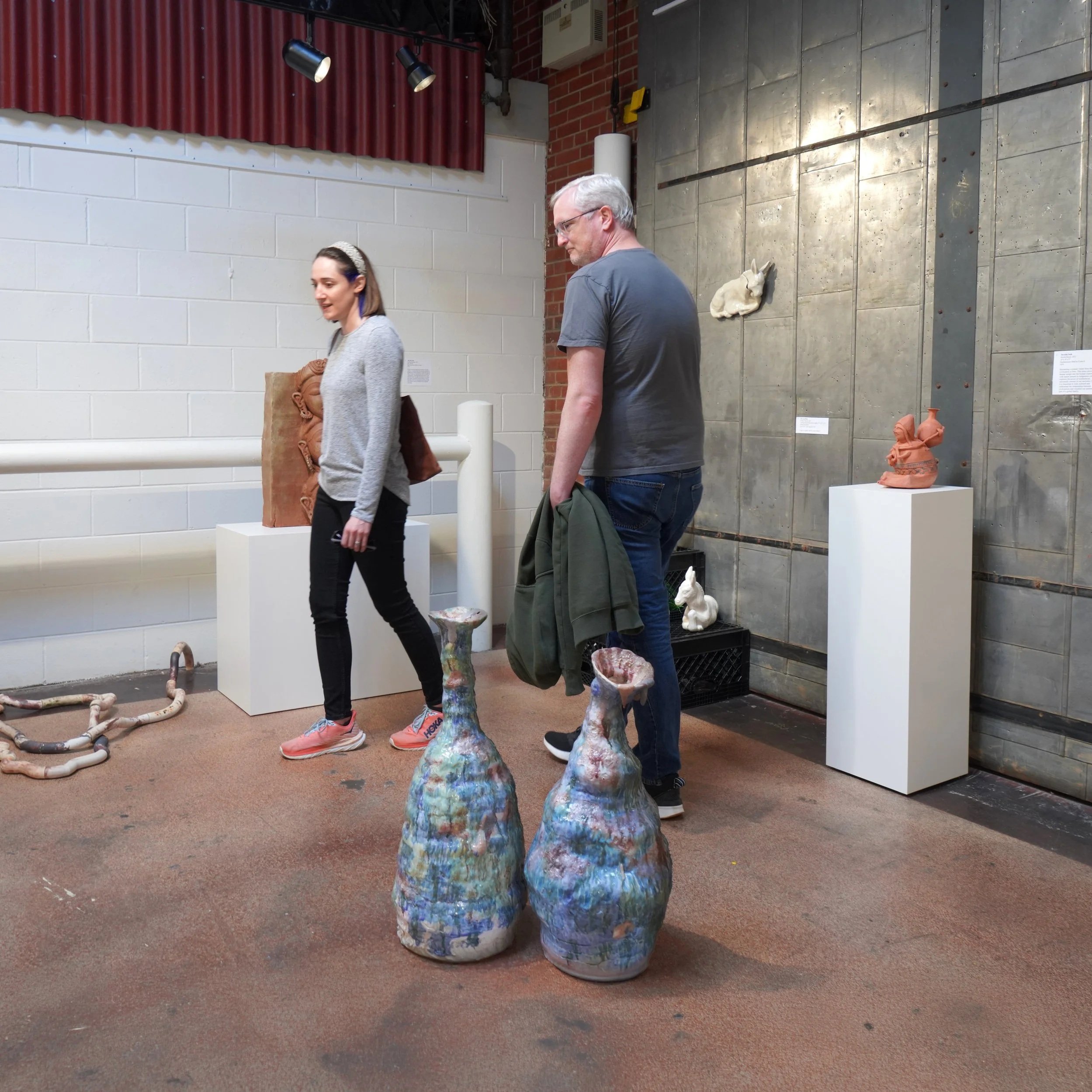 People viewing colorful ceramic sculptures of vases in an art gallery with brick and concrete walls.