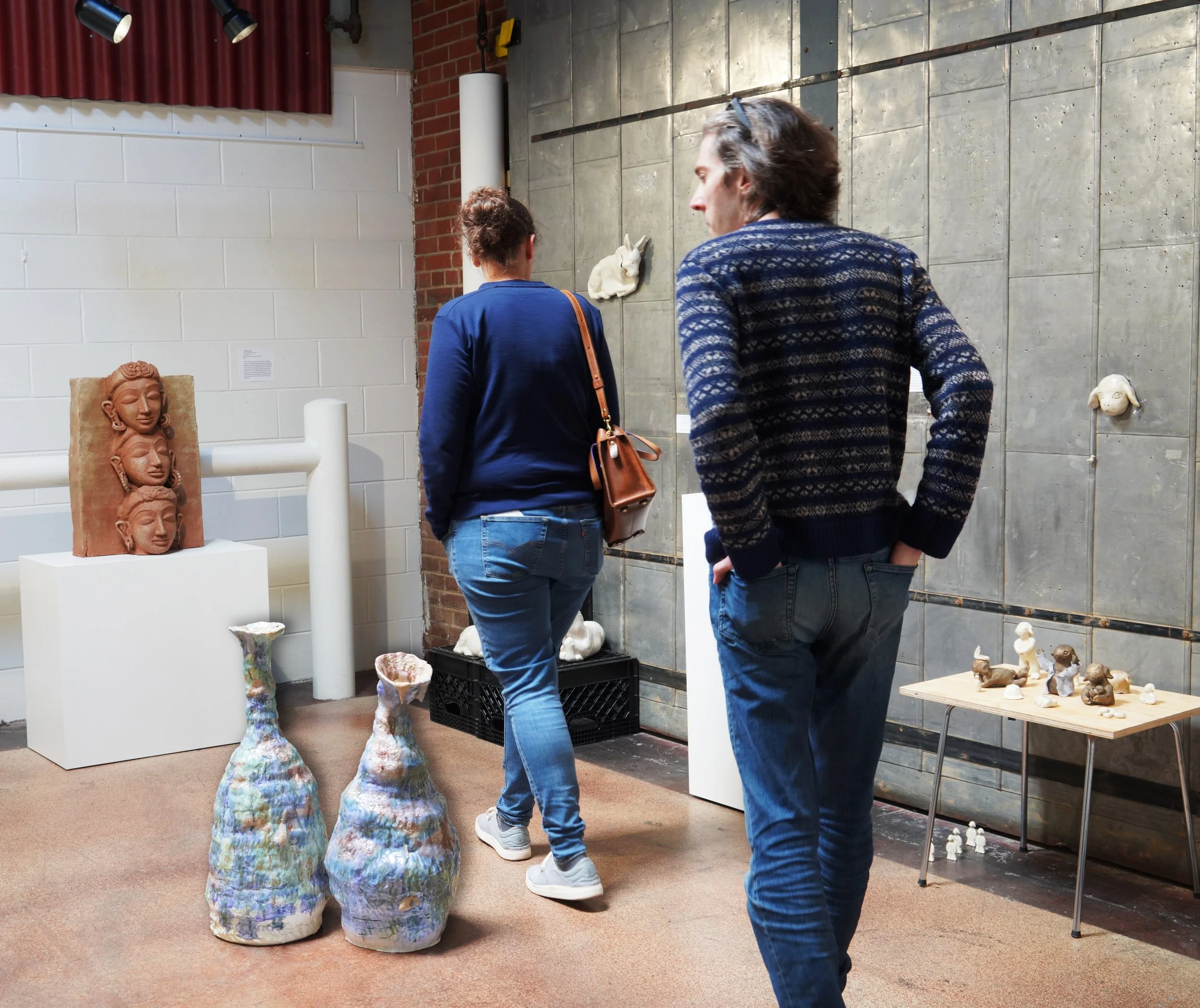 Two people are observing ceramic art sculptures in an art gallery or studio. The sculptures include a large face with intricate earrings and multiple smaller sculptures on a table. The background features a brick wall and various textures on the wall
