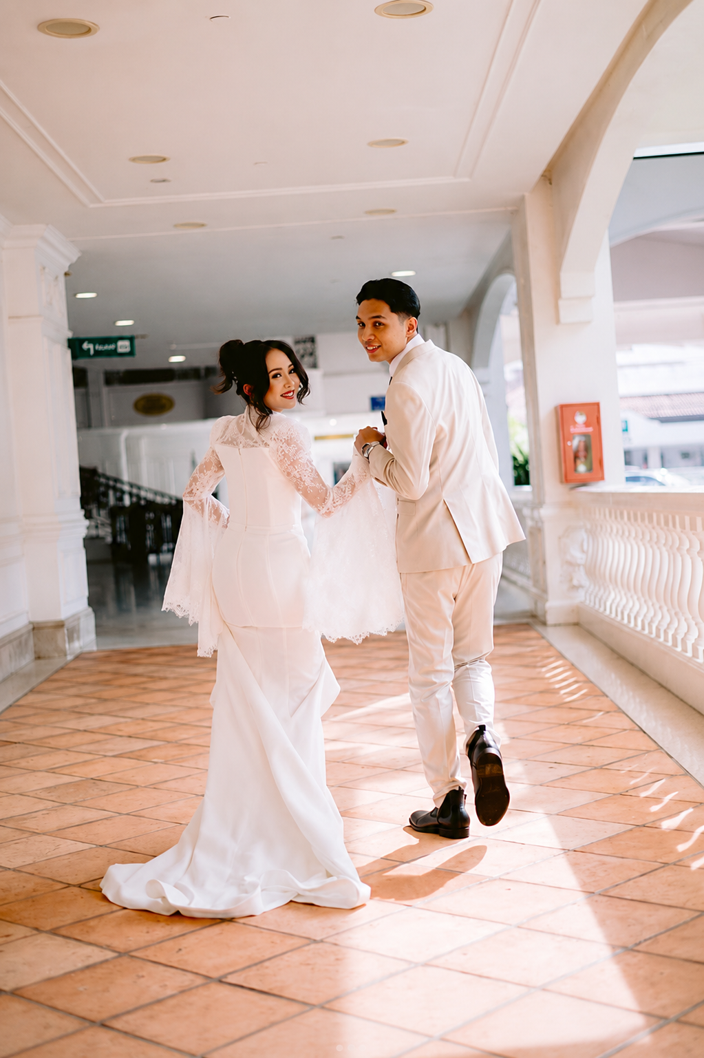 A bride and groom holding hands and walking through a bright indoor corridor, both dressed in elegant white wedding attire.