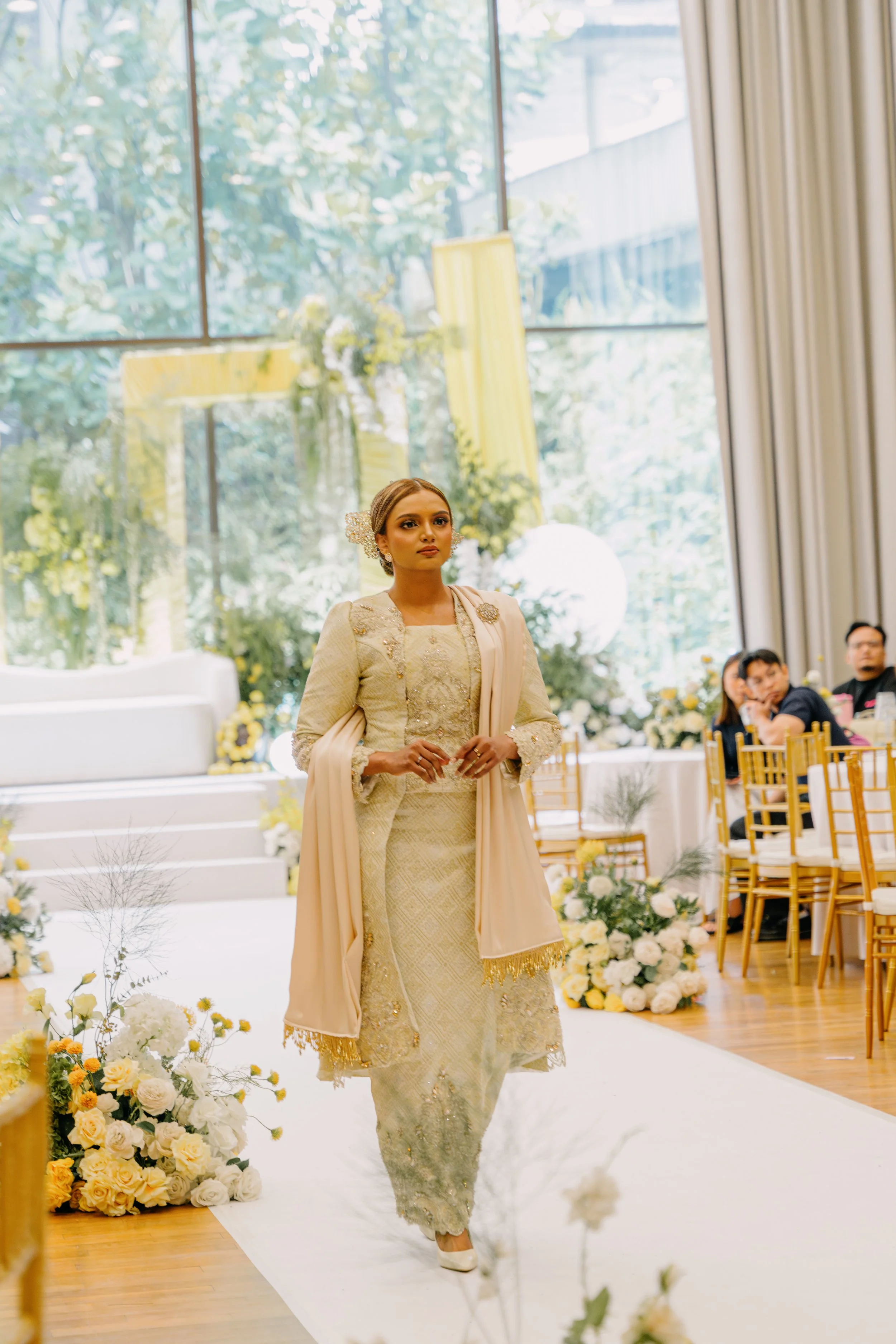 A woman dressed in traditional attire walking down an aisle decorated with flowers at a wedding or formal event in a bright, elegant venue with large windows.