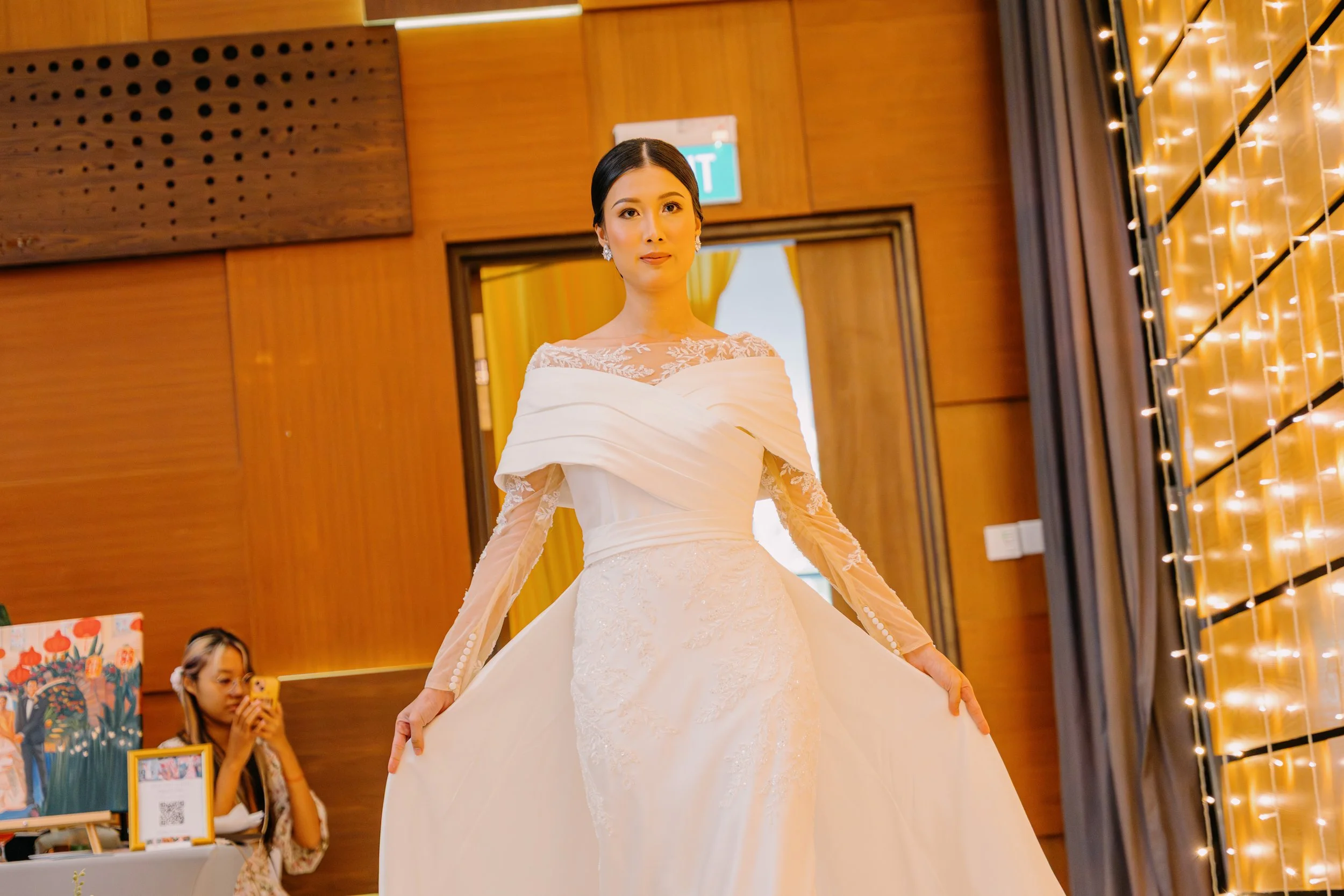 A woman in a wedding dress standing indoors with warm lighting and wooden walls, while a young girl with a yellow phone stands nearby.