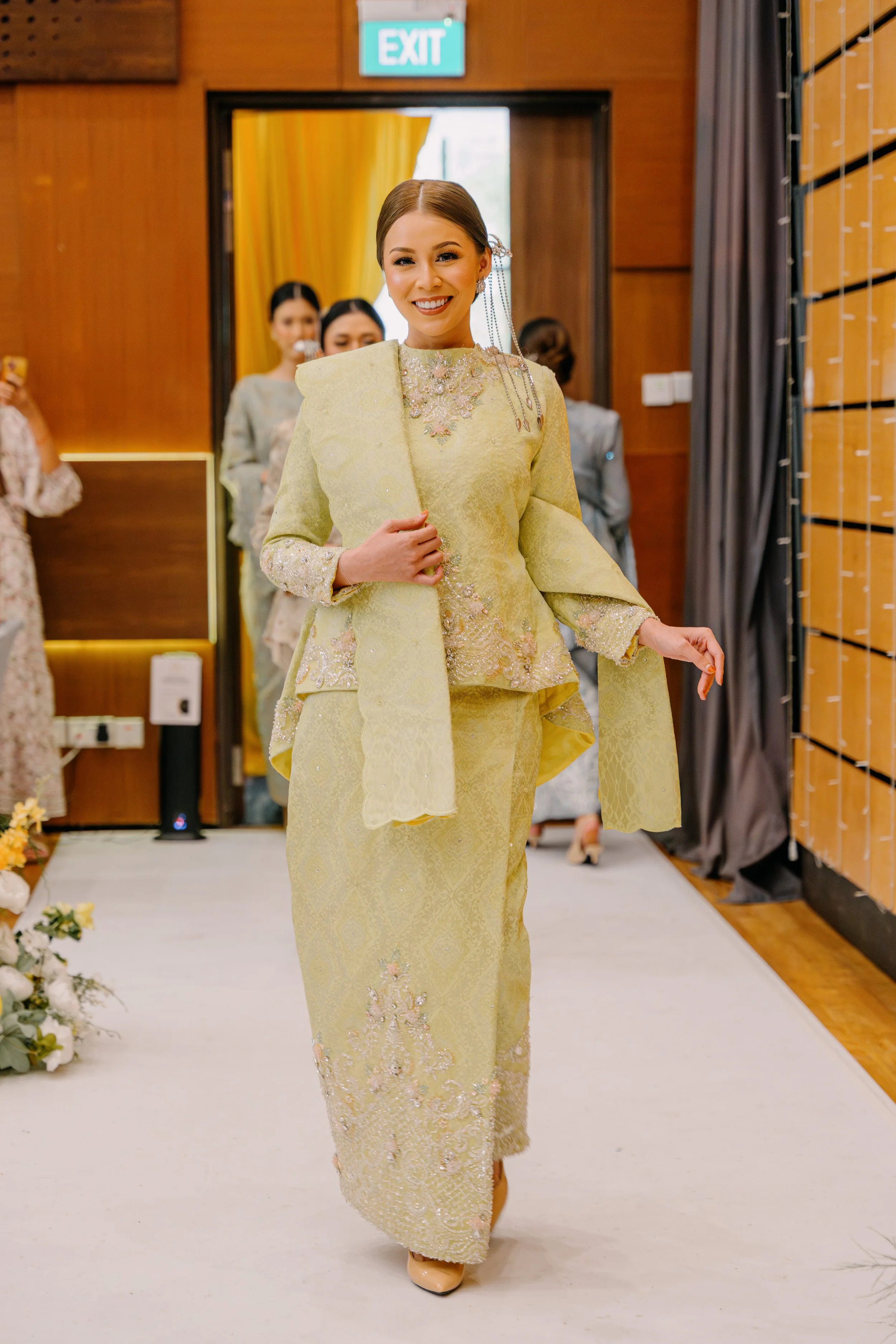 A woman wearing a traditional yellow dress with floral embroidery, smiling as she walks on a white aisle. People are visible in the background, with a wooden wall and an exit sign overhead.