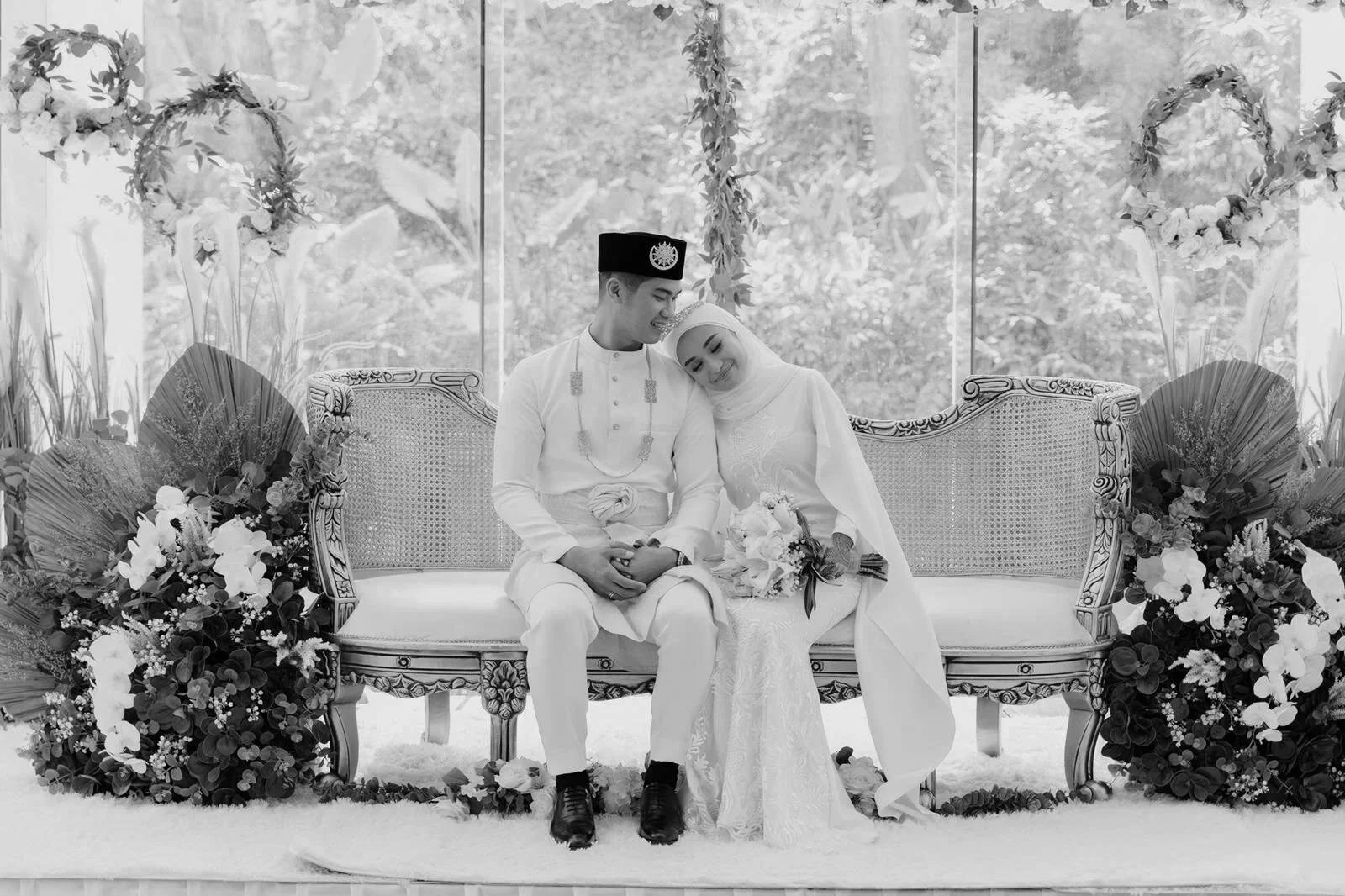 A newlywed couple in traditional Malay wedding attire sitting on a decorated sofa, smiling and leaning close to each other, surrounded by floral arrangements in an elegant setting.