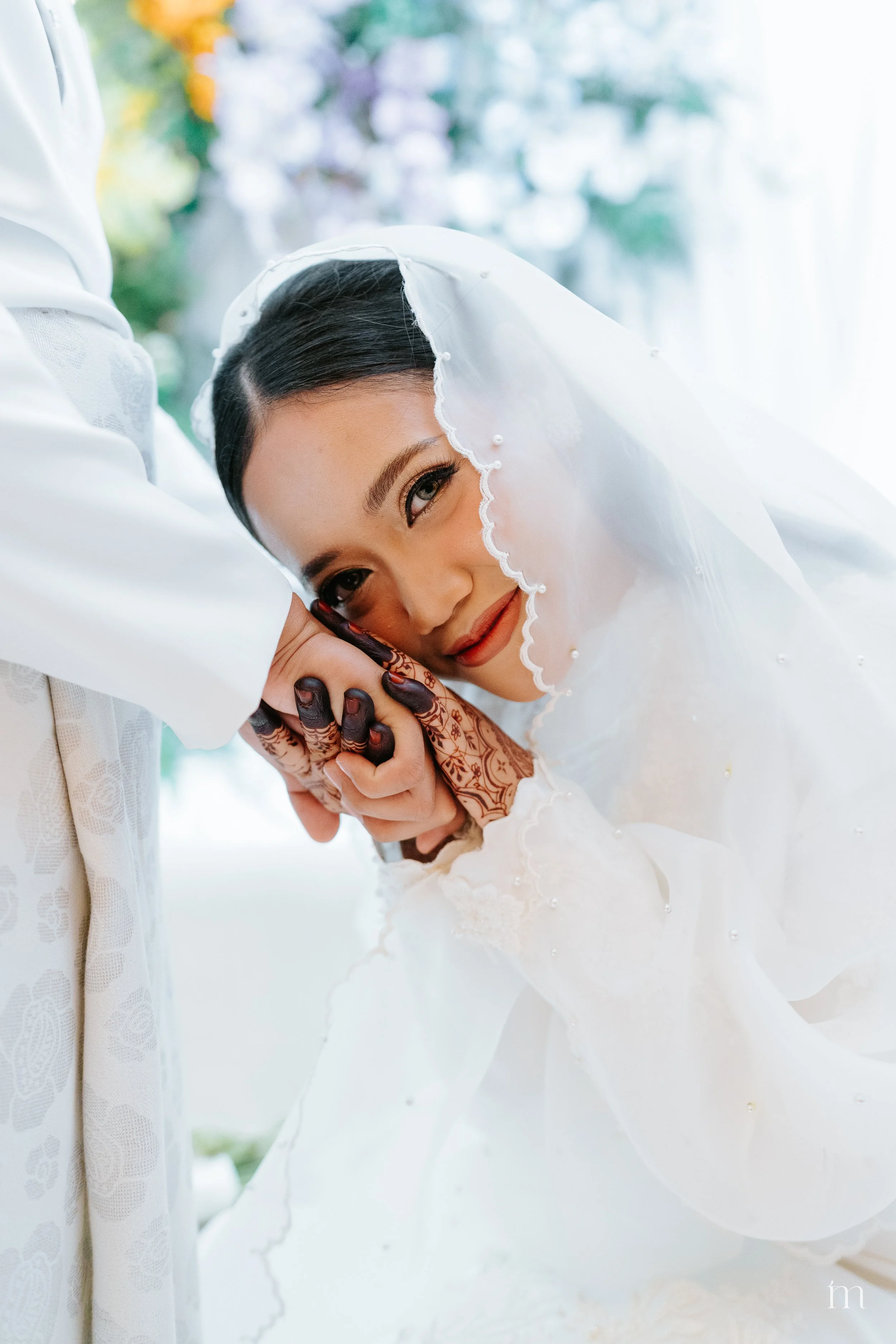 A bride with dark hair and makeup smiling softly, resting her head on her clasped hands, which are decorated with intricate henna designs, underneath a white veil with pearl accents, during a wedding ceremony.