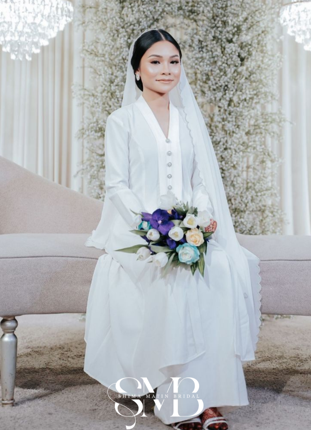 A woman dressed in white, sitting on a beige sofa, holding a bouquet of white roses, purple flowers, and greenery, in front of a background of white flowers and chandeliers.