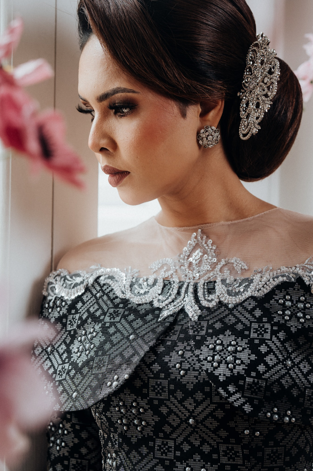 A woman with styled dark brown hair in a low bun, wearing intricate pearl and crystal earrings and a black garment with white lace and bead embroidery, looking to the side with a serene expression.