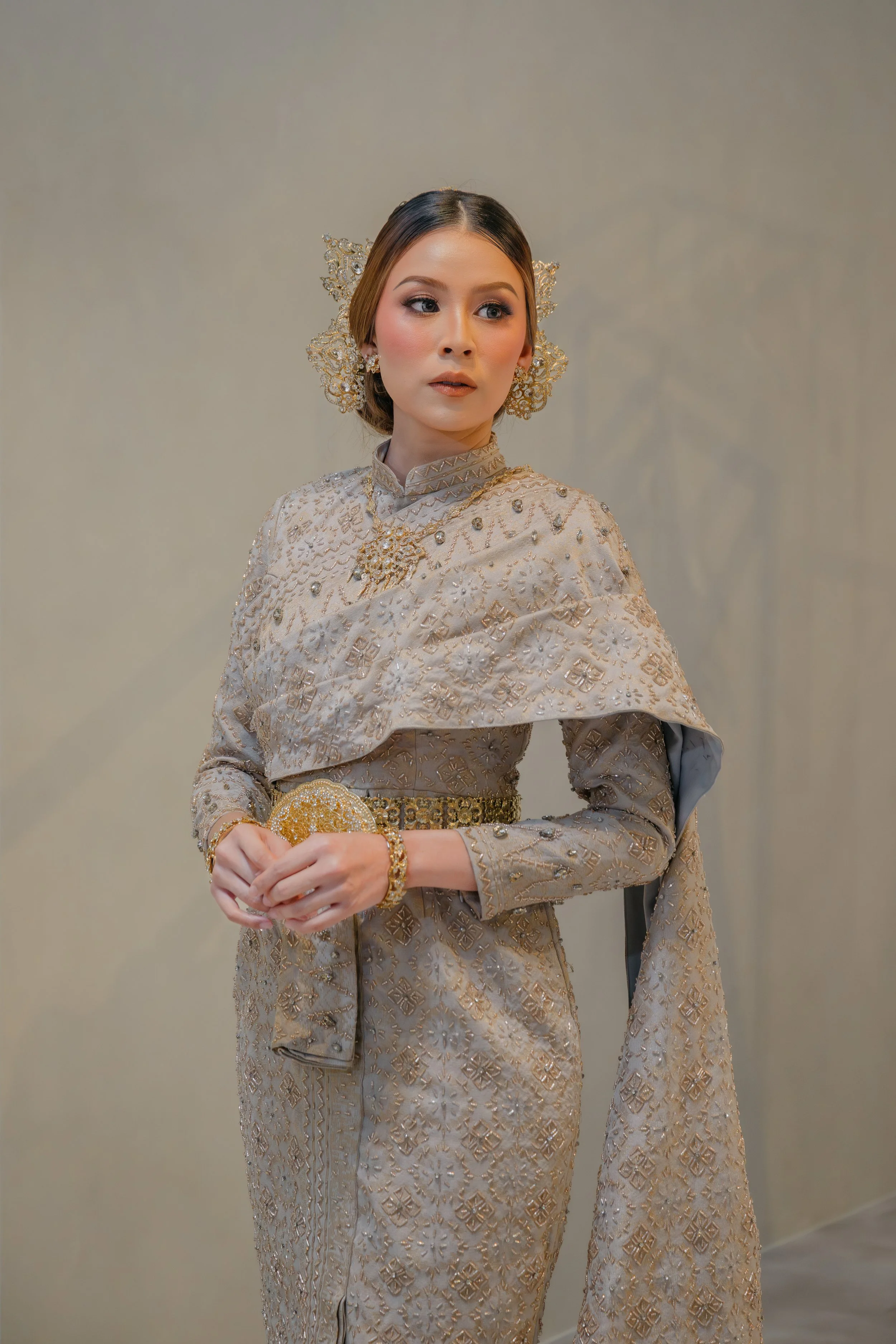 A woman dressed in traditional Thai attire, with intricate gold embellishments, ornamented jewelry, and ornate headpieces, holding a gold clutch.
