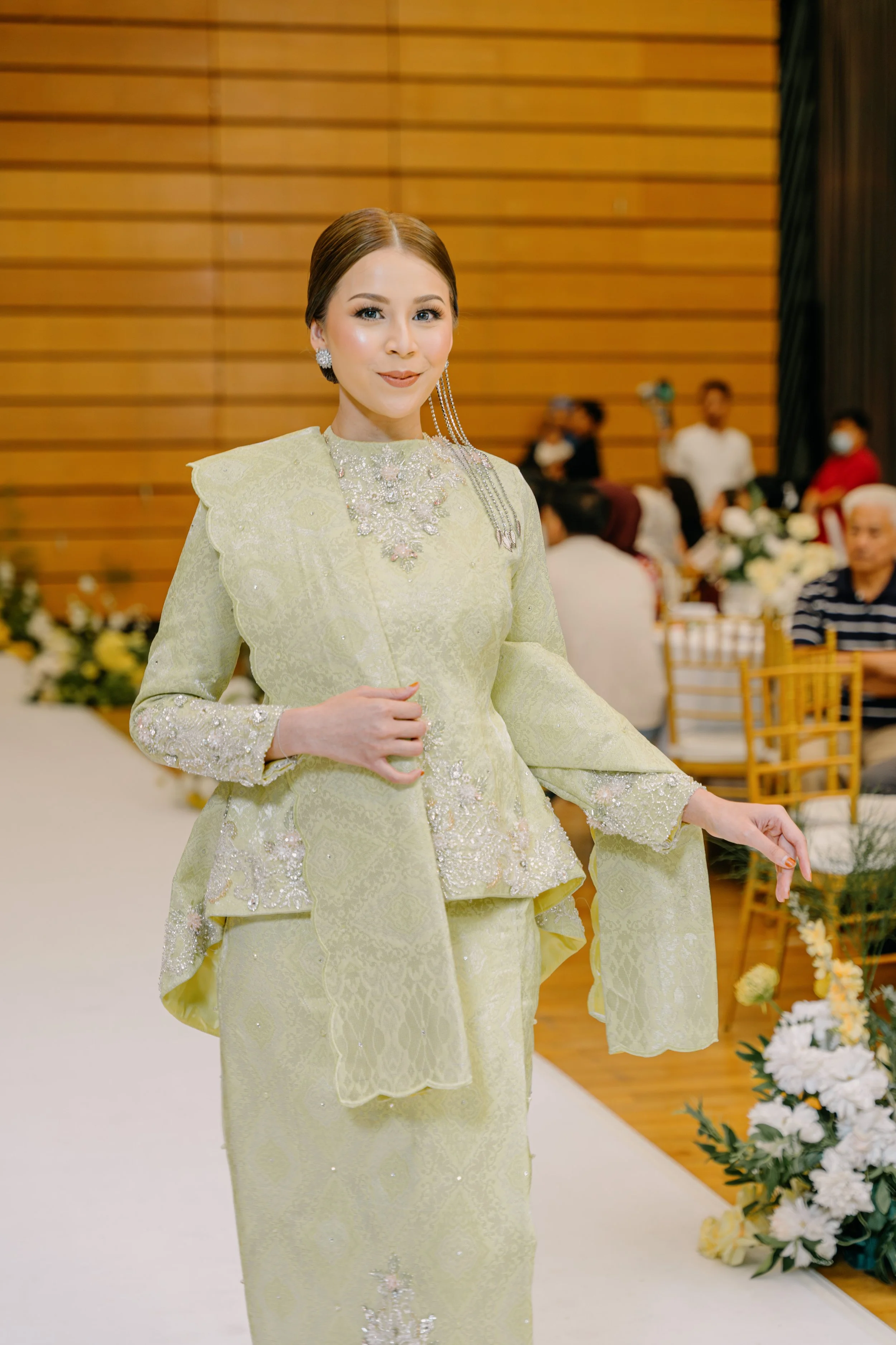 A woman dressed in traditional yellow attire with detailed silver embroidery, standing in a decorated indoor setting with people and floral arrangements in the background.