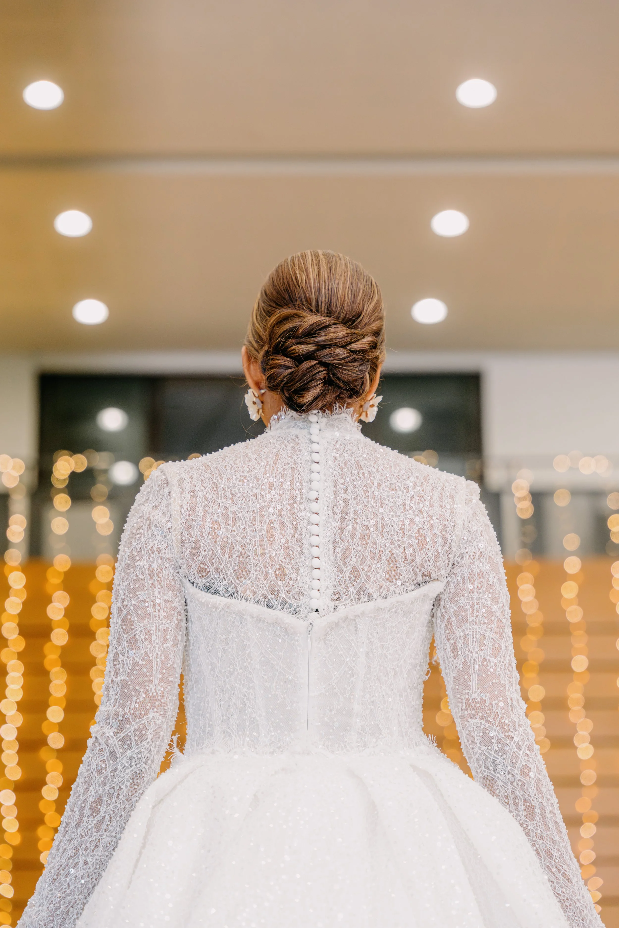 Back of a bride in a lace wedding gown with buttons down the back, dark brown hair styled in an elegant updo, standing in front of blurred warm lights and modern ceiling lights.