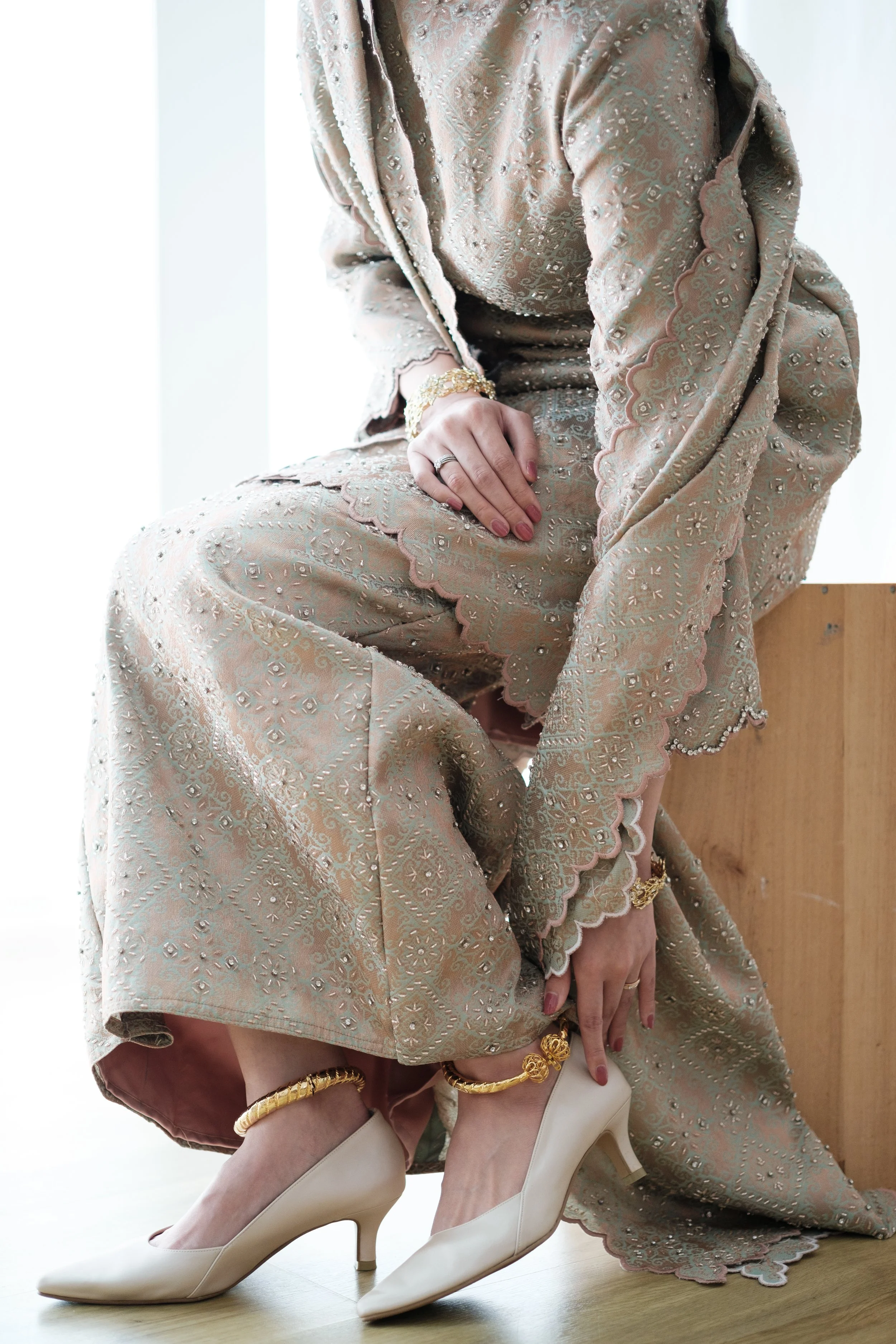 Close-up of a woman in elegant beige lace embroidered dress with scalloped edges, wearing cream heels and gold jewelry, sitting on a wooden stool on a light wooden floor, with a white background.