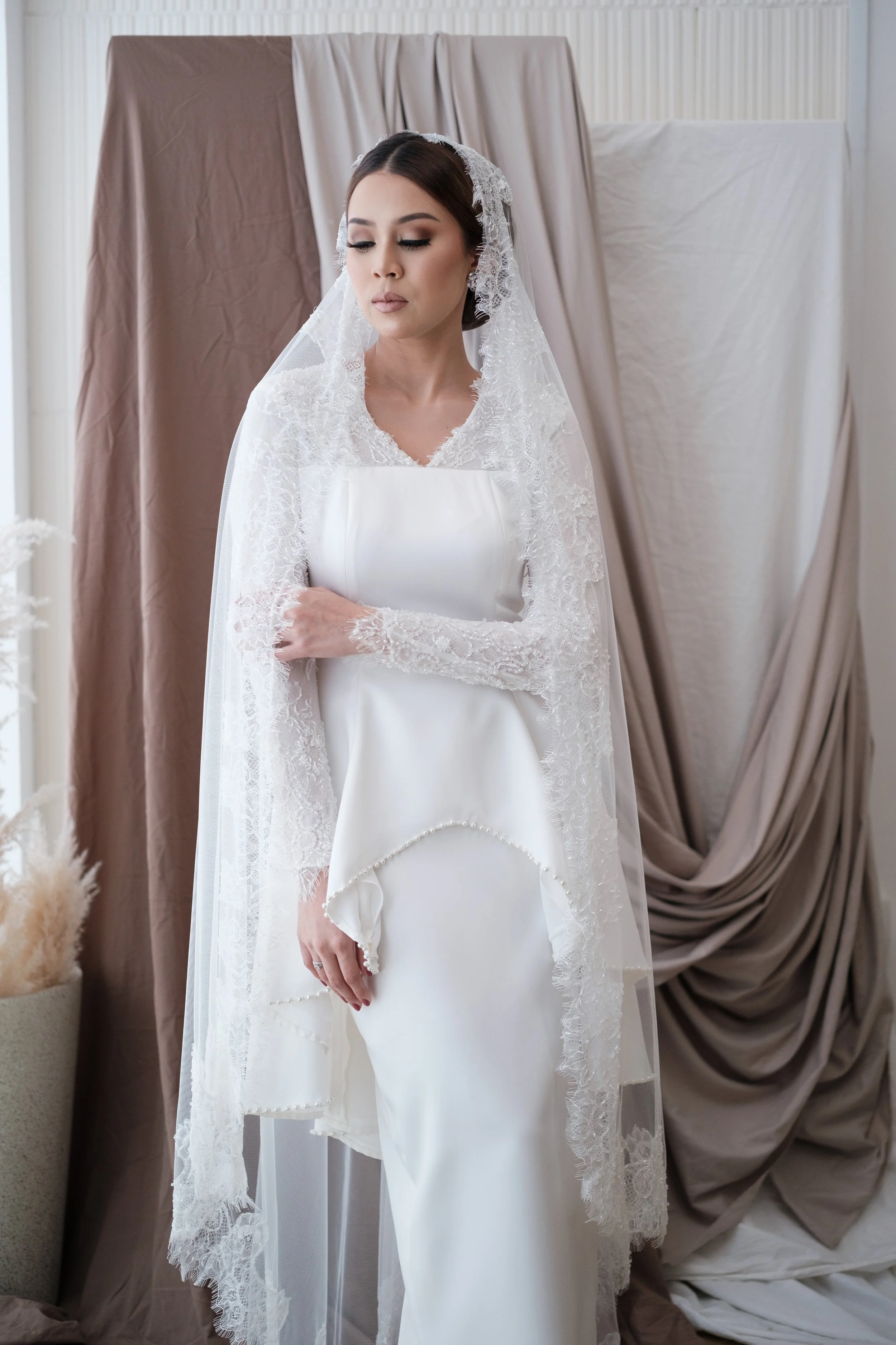 A woman in a white wedding dress with lace accents, standing indoors with her eyes closed and arms crossed, behind a backdrop of beige and white drapes.