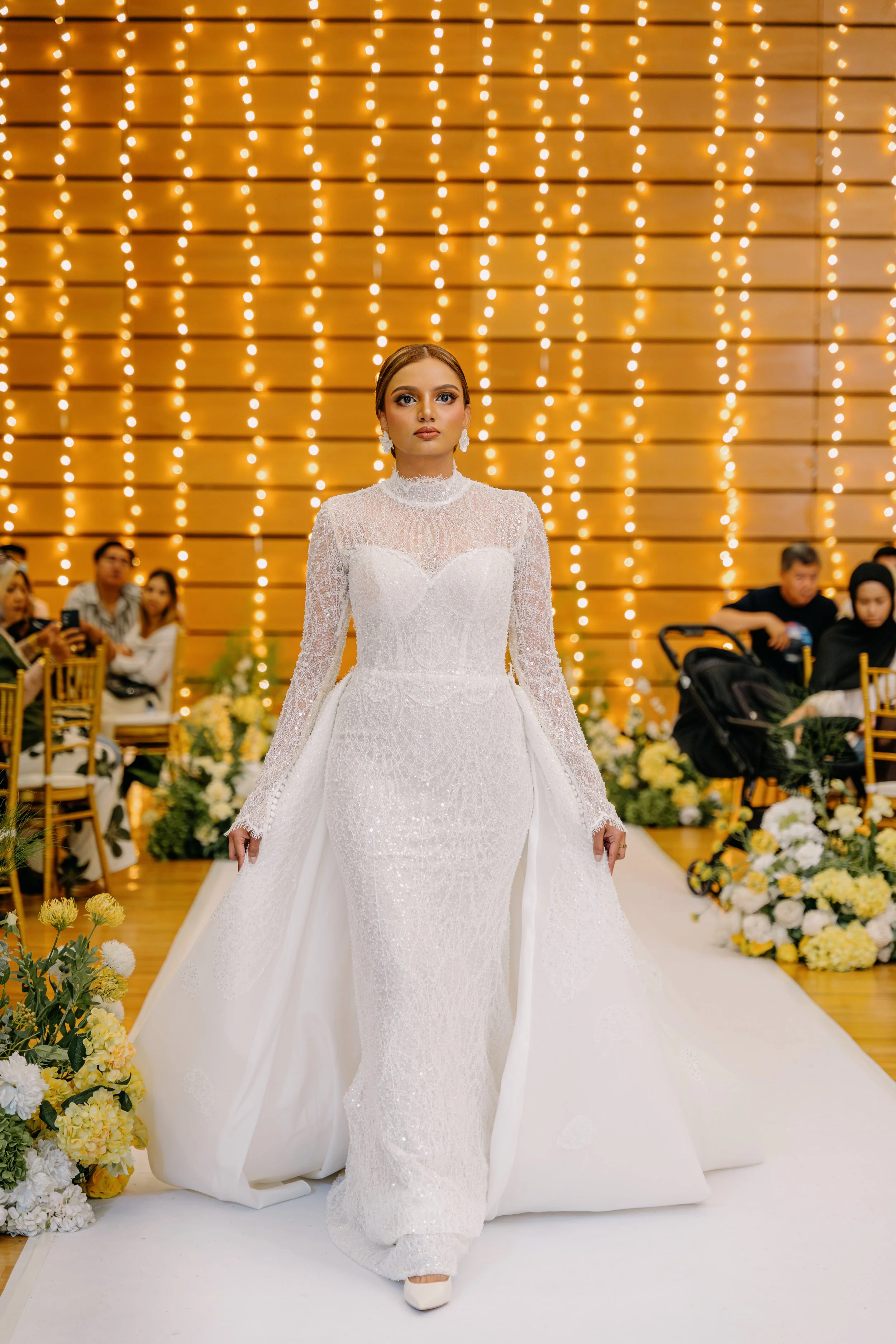 A woman in an elegant white wedding gown walking down the aisle at a wedding ceremony, with guests seated on either side and a wooden backdrop decorated with yellow and white flowers.