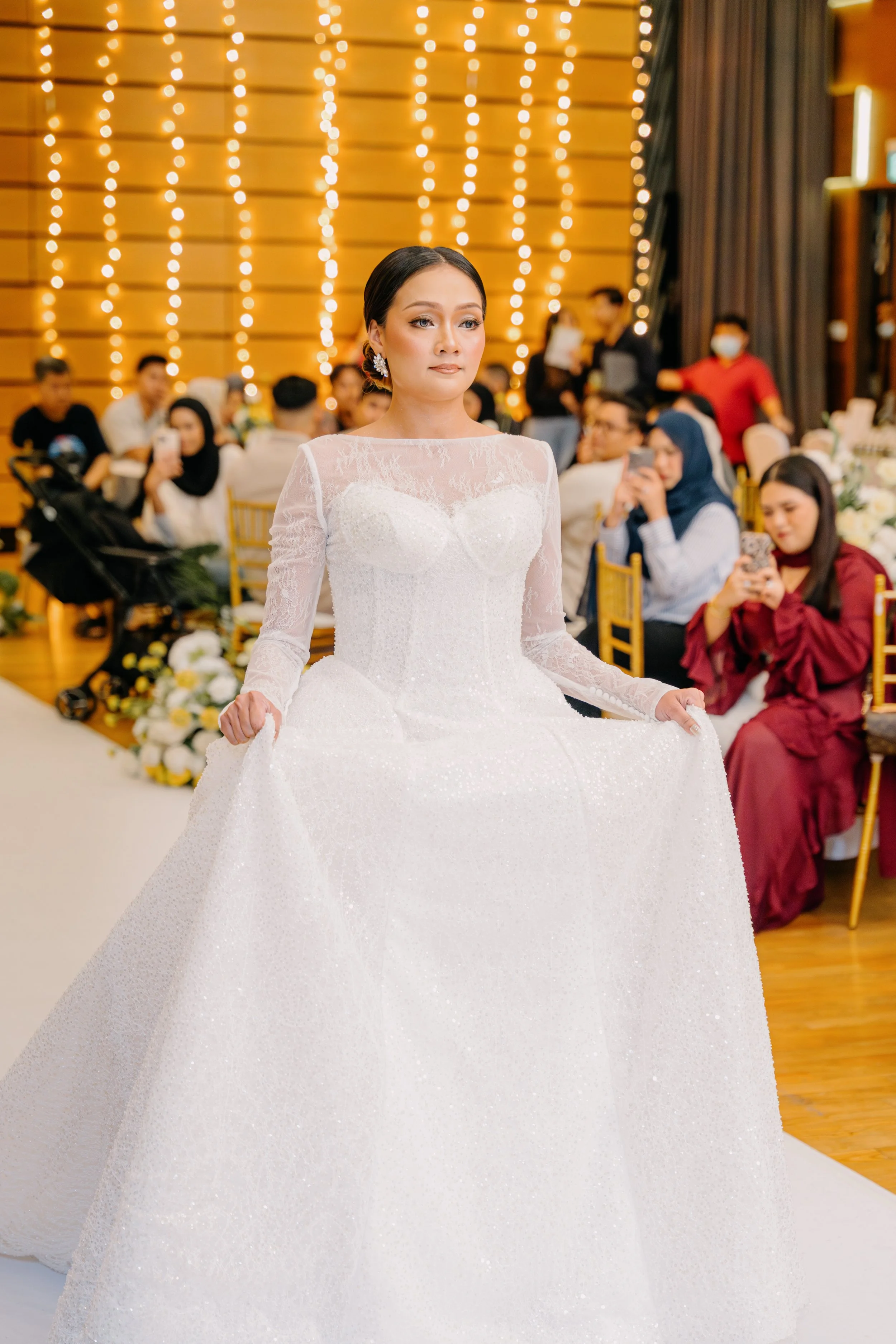 A bride in a white wedding gown with lace sleeves, sitting at a wedding reception with guests in the background, some taking photos and videos, in a decorated venue with warm lighting and wooden walls.