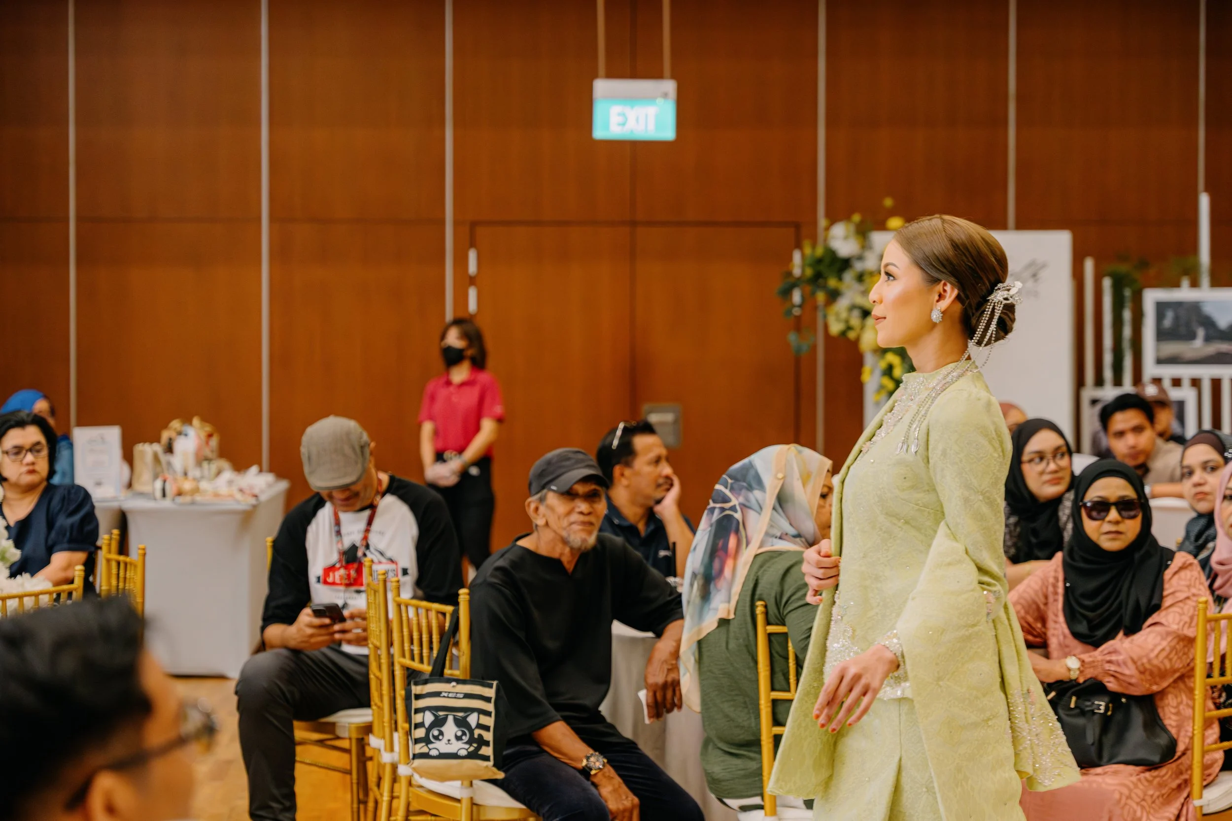 A woman dressed in traditional attire standing in a room with people seated in chairs, some looking at their phones, and a waitress in a pink uniform in the background.