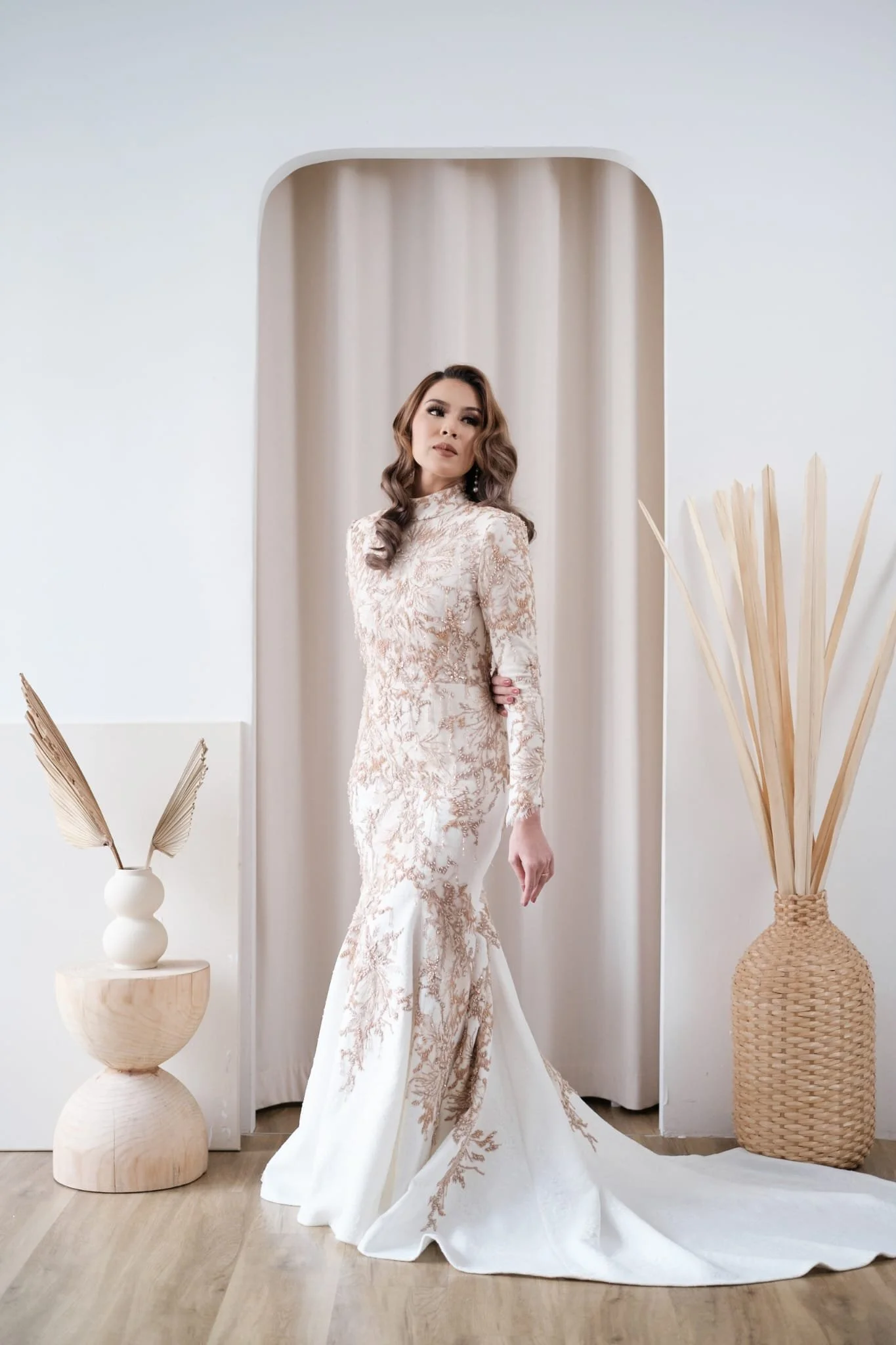 A woman in an elegant white and gold lace wedding dress standing in a minimalist room with white walls and wooden flooring, decorated with neutral-colored vases and dried plant arrangements.