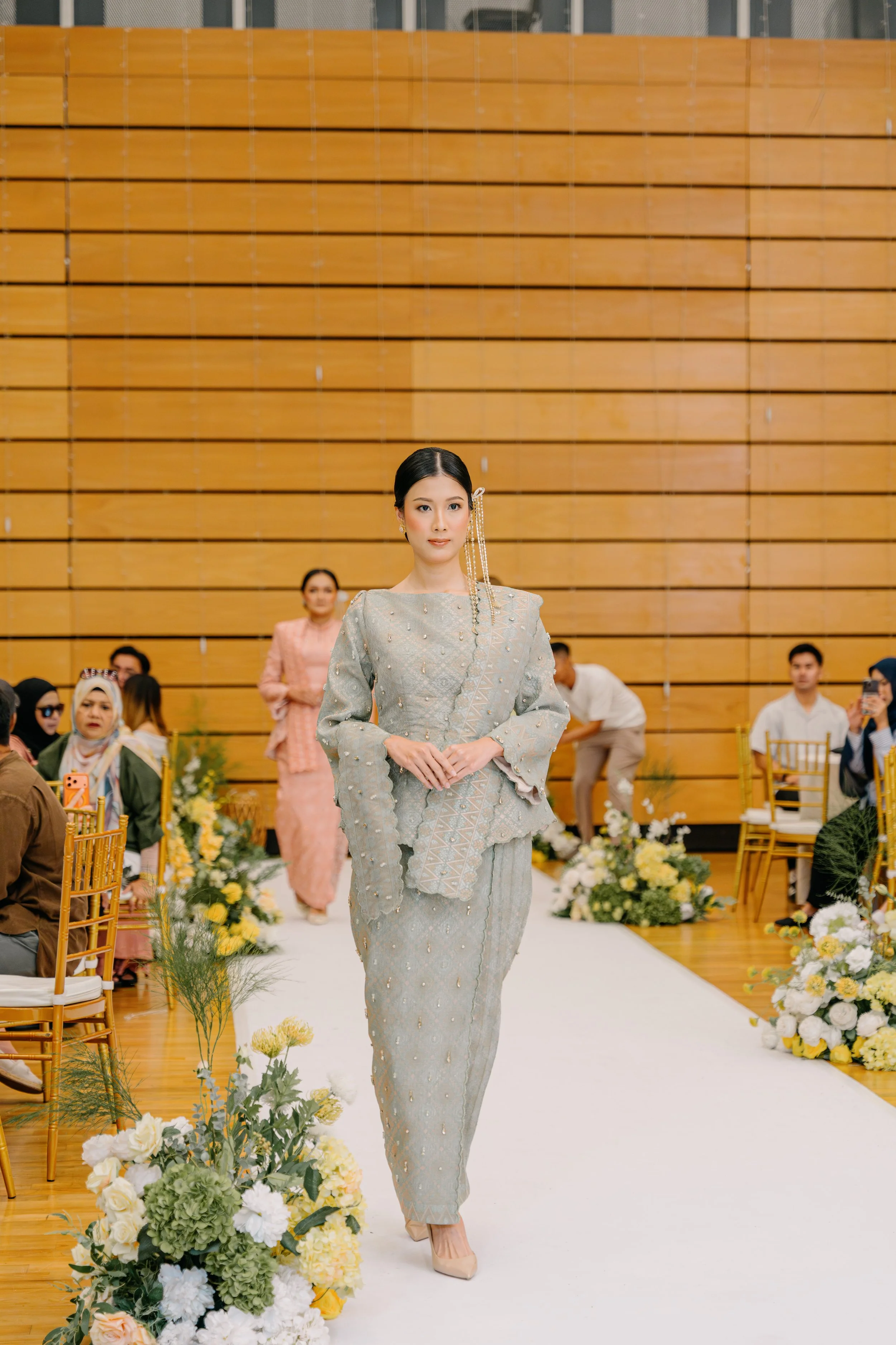 A woman in traditional dress walking down an aisle at a formal event, surrounded by floral arrangements.