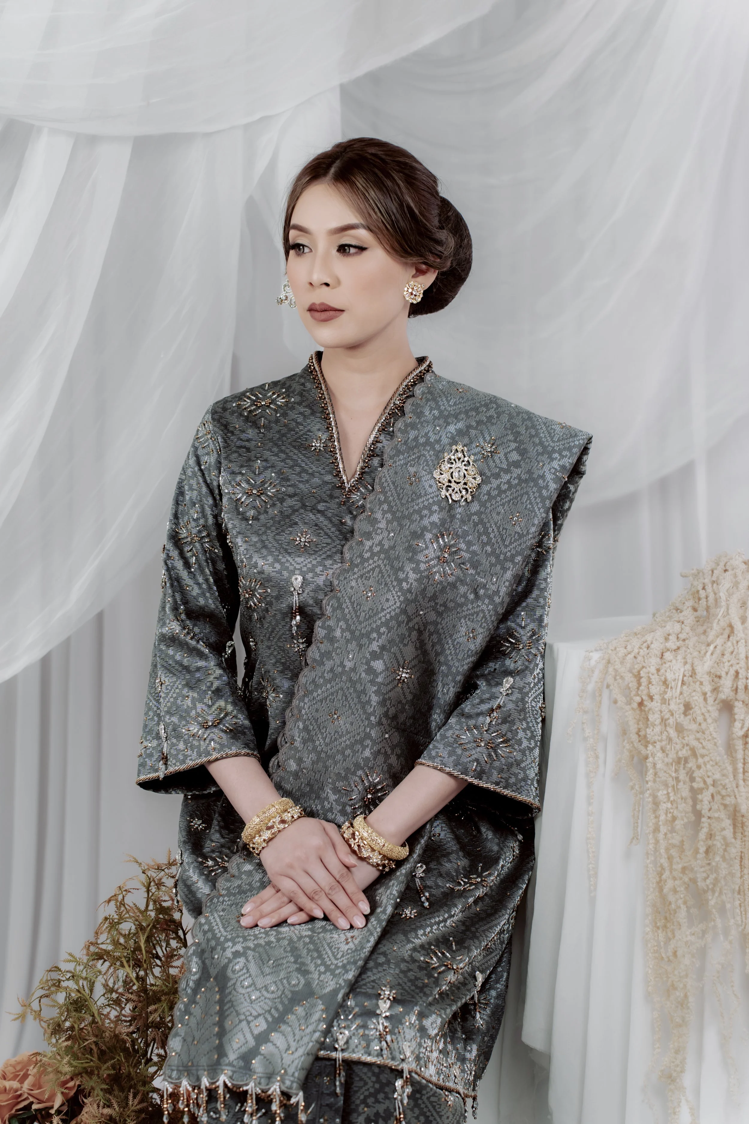 A woman dressed in a traditional patterned gray and silver outfit with gold jewelry, seated with her hands resting on her lap, against a backdrop of sheer white drapes and some beige decorative elements to her right and bottom left.