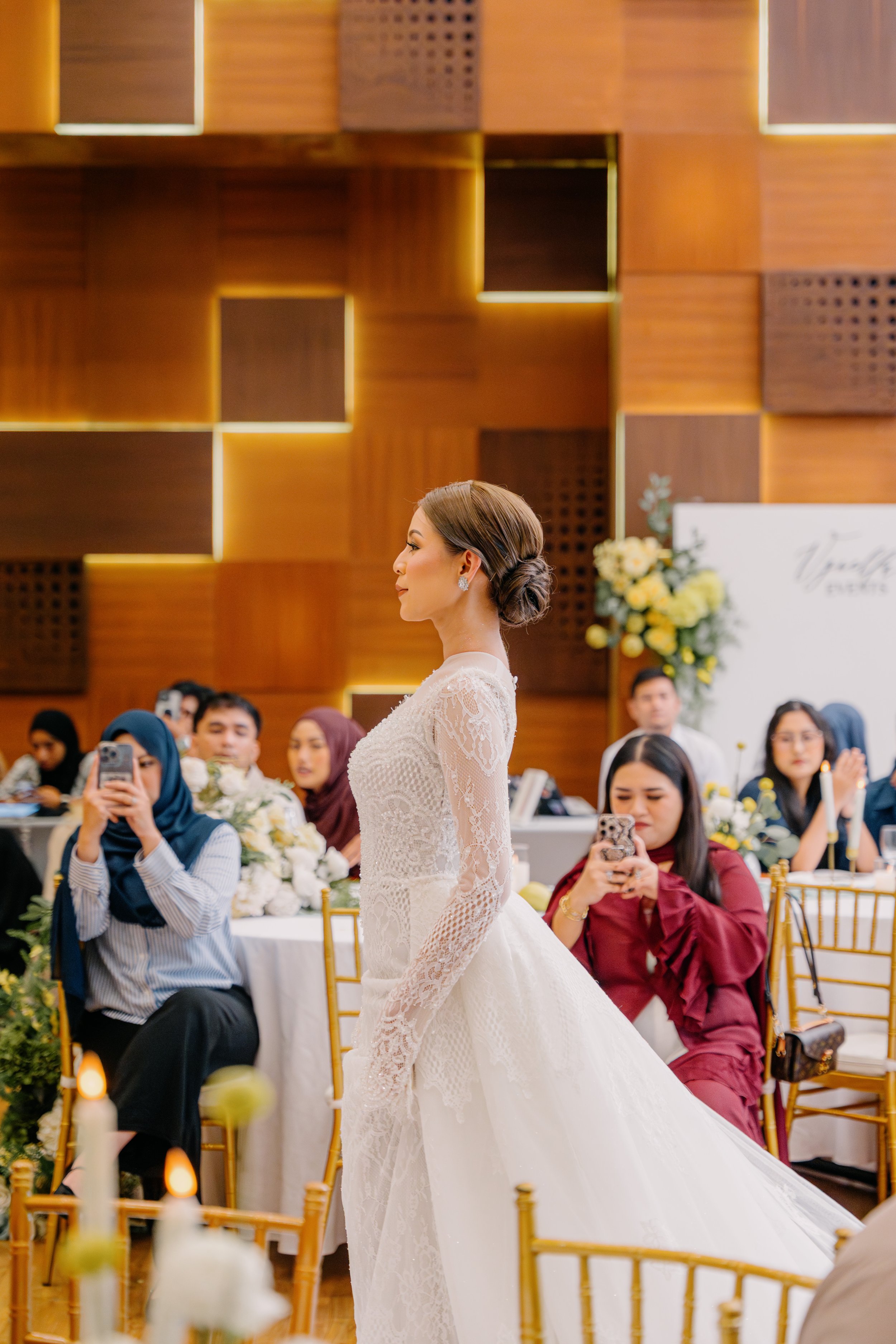 A woman in a white wedding dress standing in an elegant wedding reception hall filled with seated guests taking photos and videos with their smartphones.
