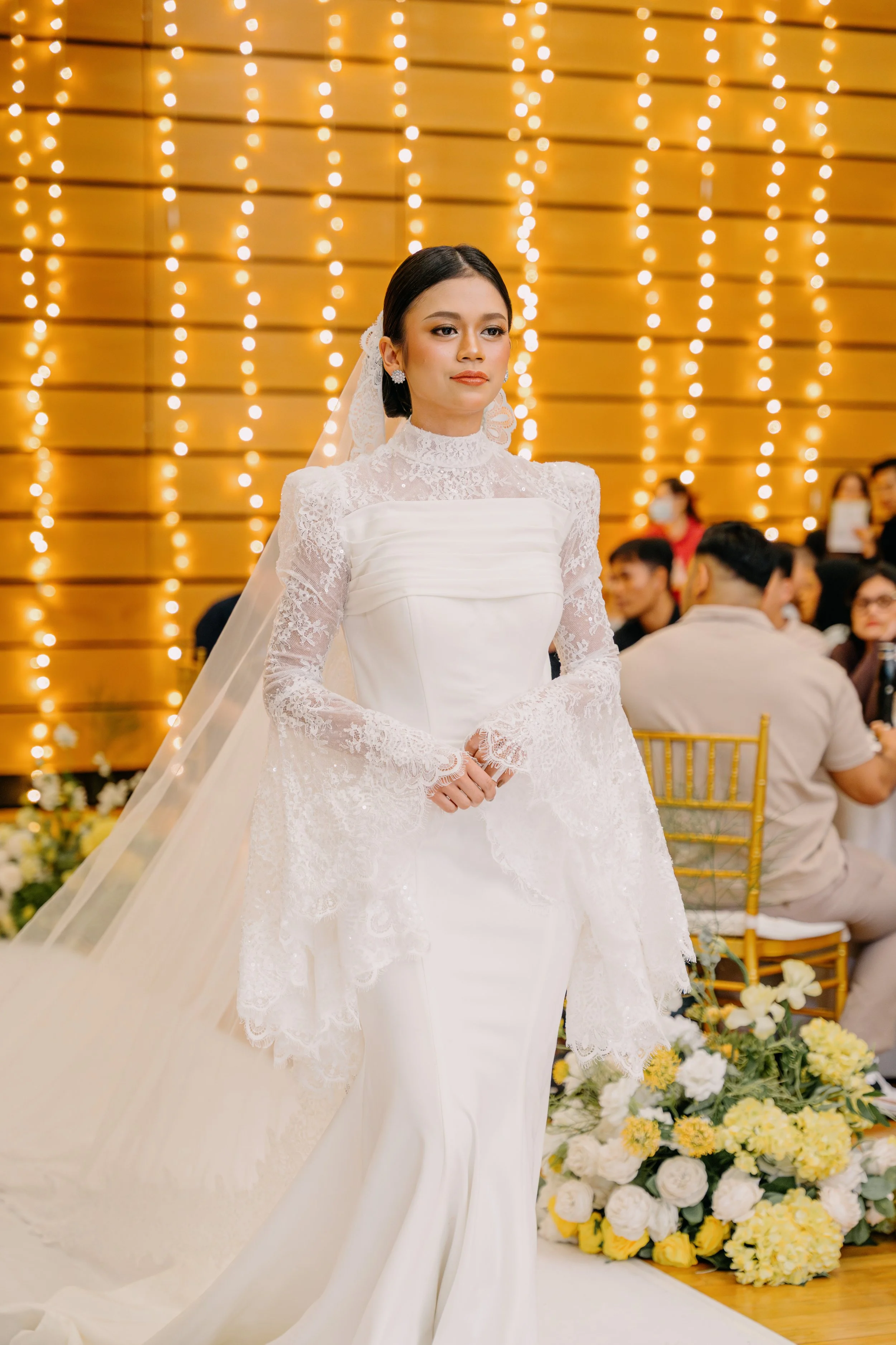 A bride in a white lace wedding dress with a veil, standing in a decorated wedding venue with warm lighting and floral arrangements.