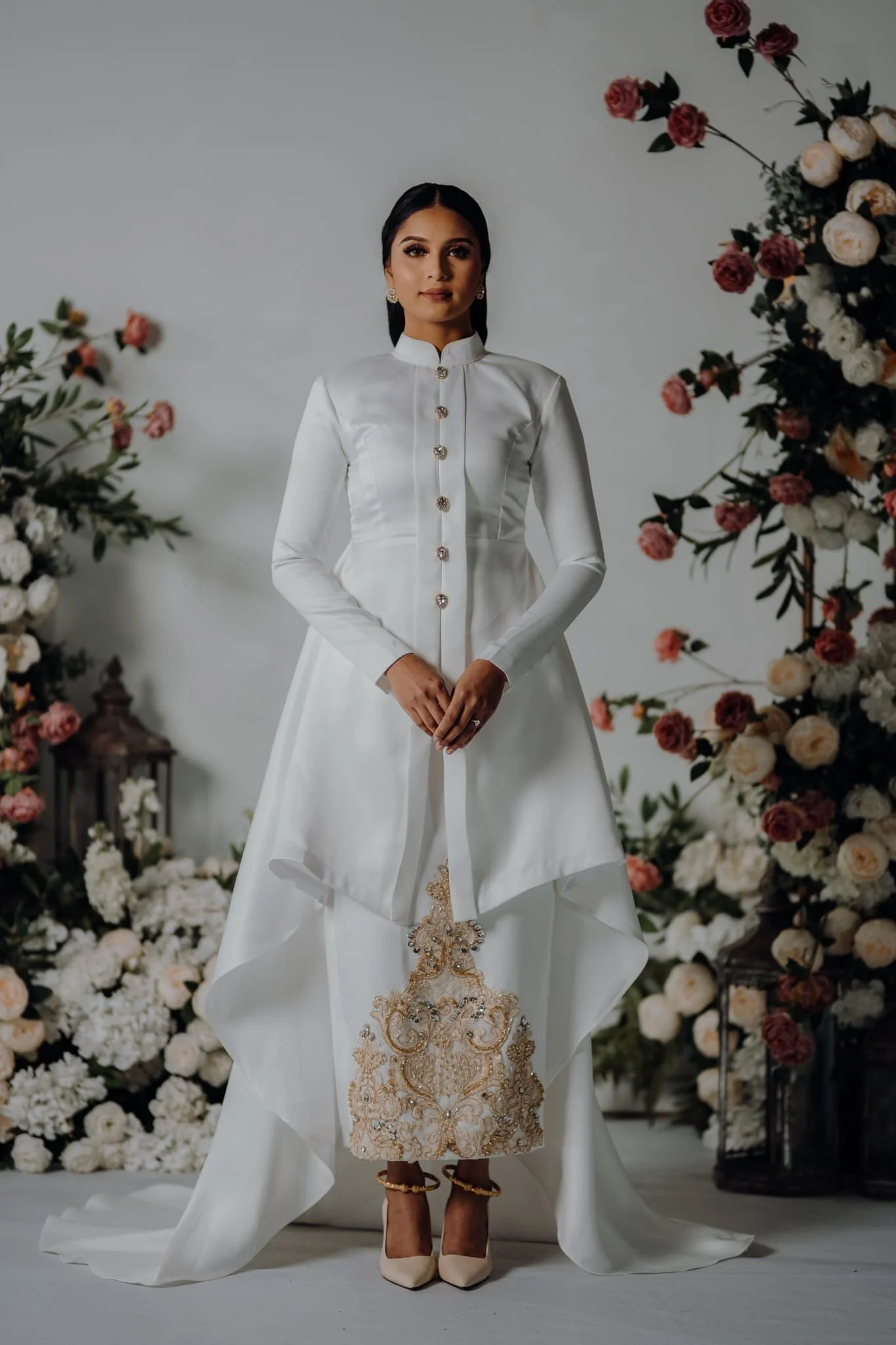 A woman in an elegant white dress with gold embroidery standing in front of a floral backdrop. The dress features long sleeves, a high collar, and ornate buttons. She is wearing beige heels and has dark hair styled back, accessorized with earrings. T