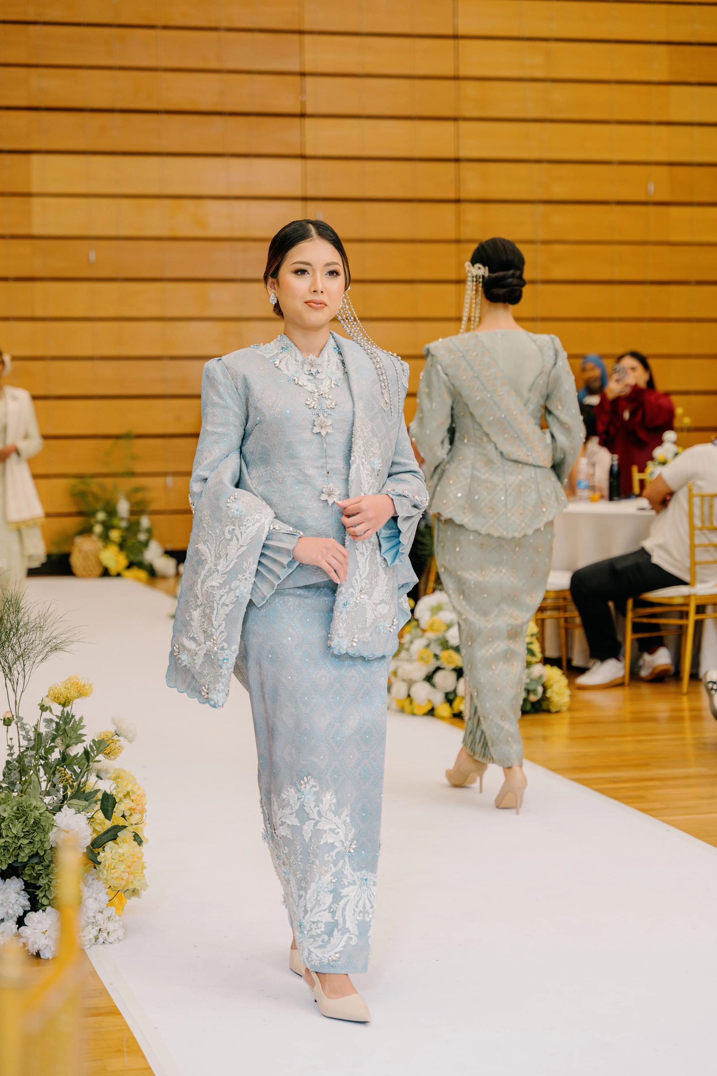 Women dressed in elegant traditional dresses participating in a fashion event or ceremony, with one walking on a white runway and others standing nearby in a venue with wooden walls.