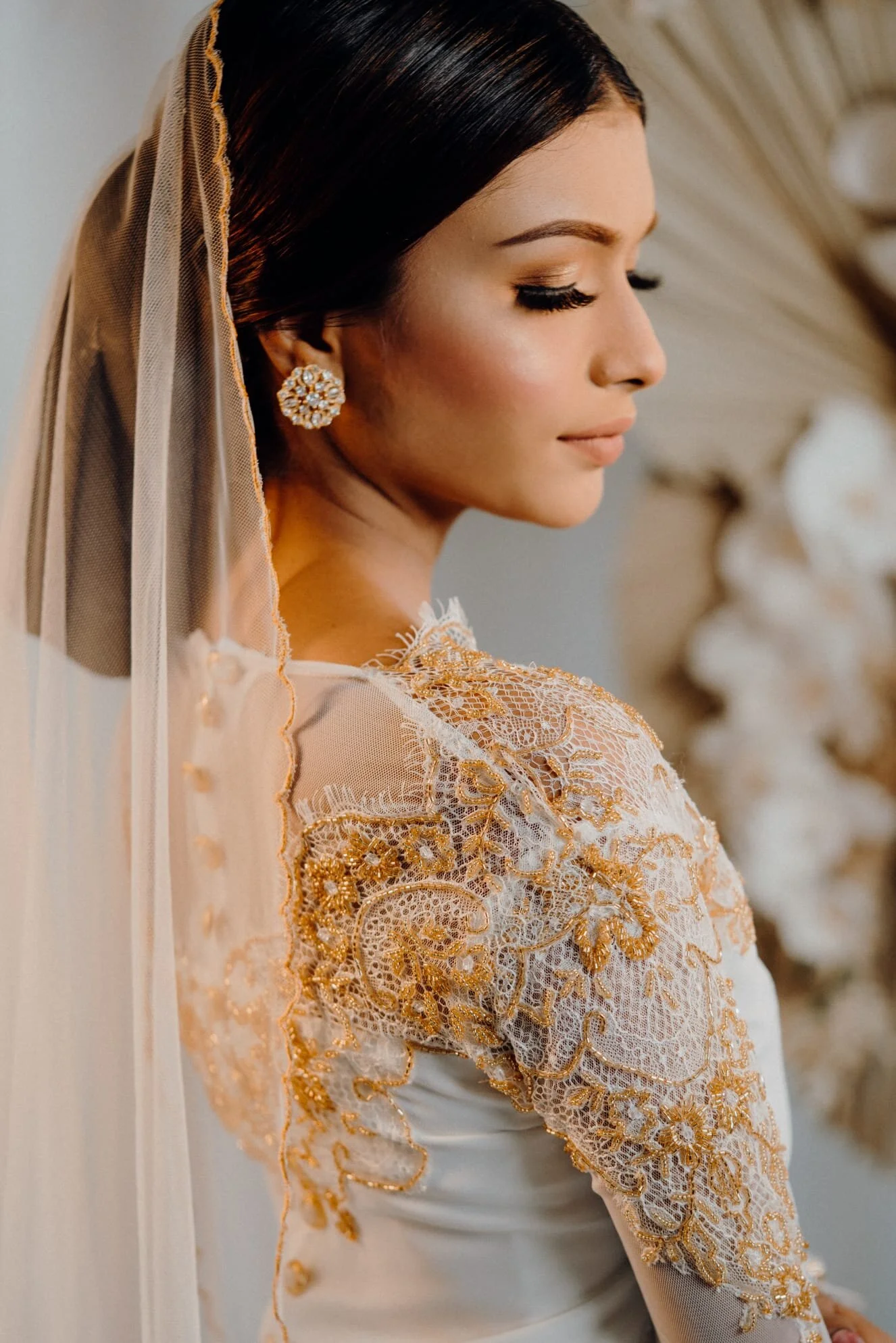 Close-up of a woman with dark hair wearing ornate gold and white lace dress and large floral-style earrings, standing against a light background.