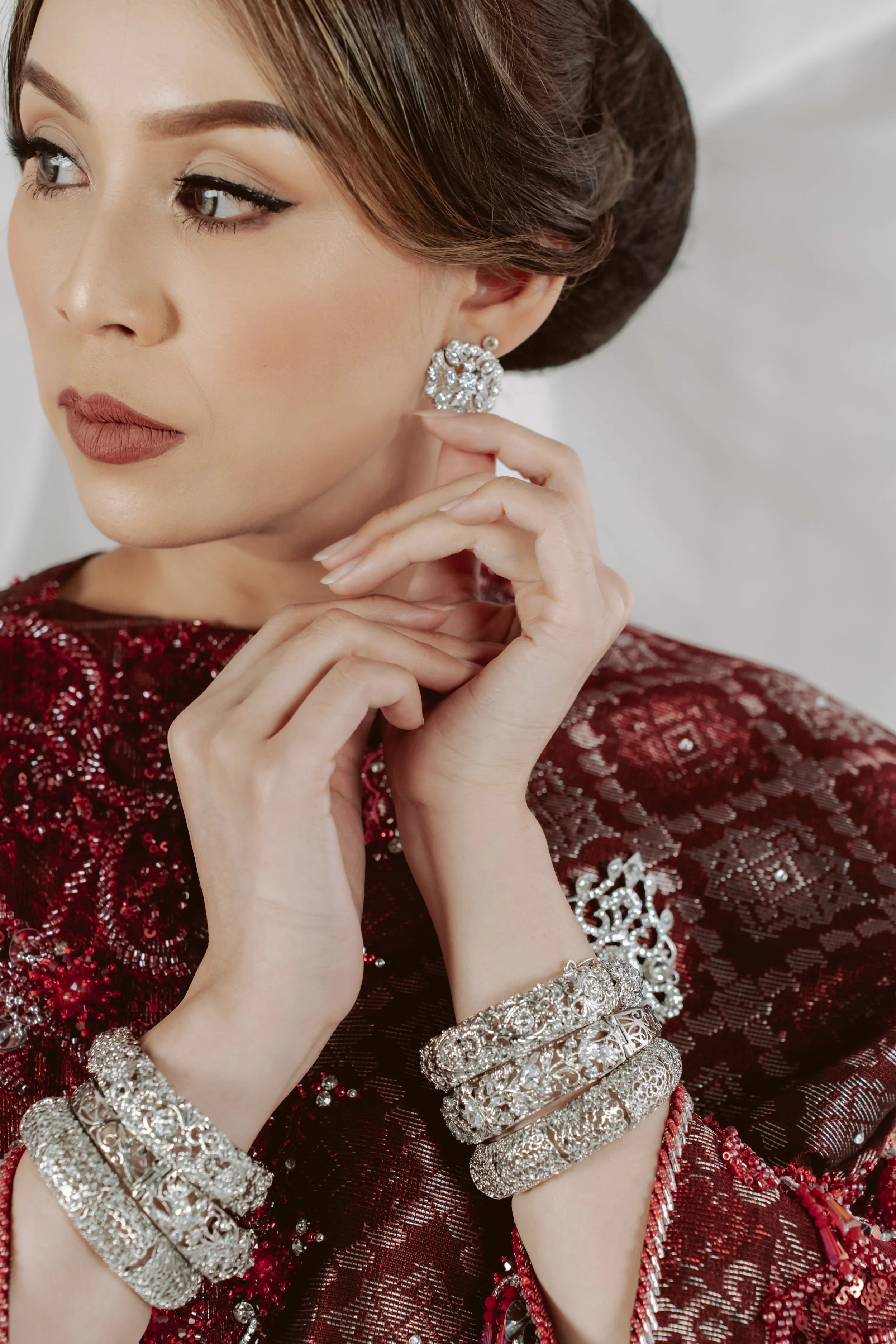 Close-up of a woman with makeup and jewelry, wearing a red and silver dress, touching her ear with her hand, which is adorned with multiple silver bracelets.