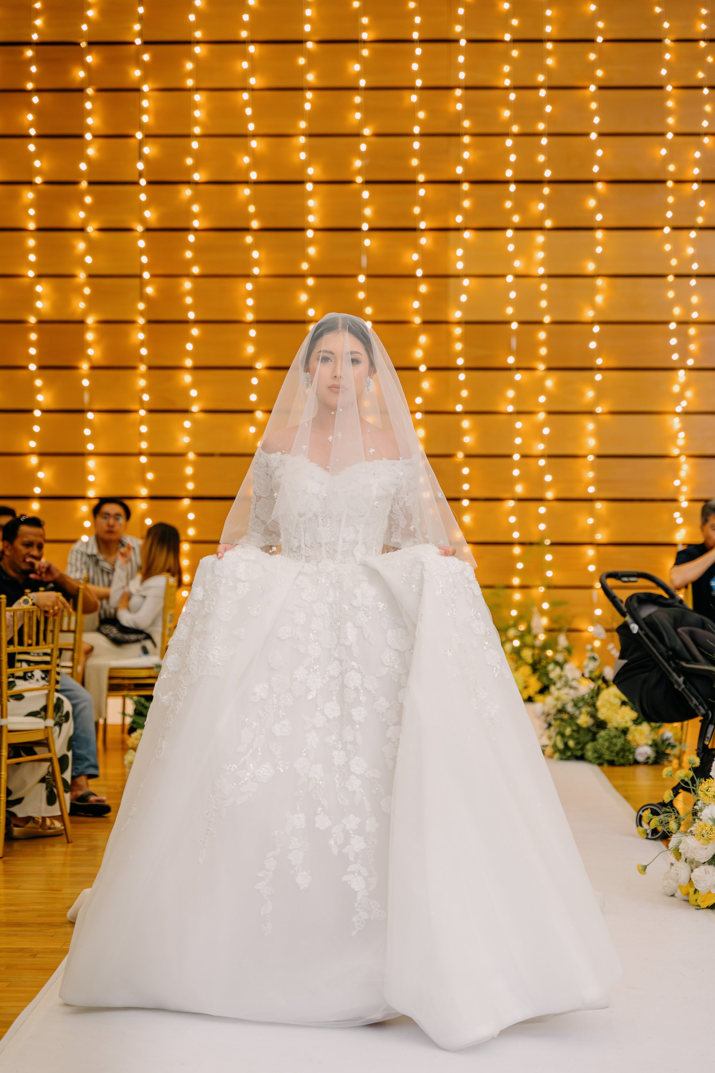 Bride in a wedding gown with a veil holding the dress, standing in front of a backdrop of warm string lights, with seated guests and floral arrangements in the background.