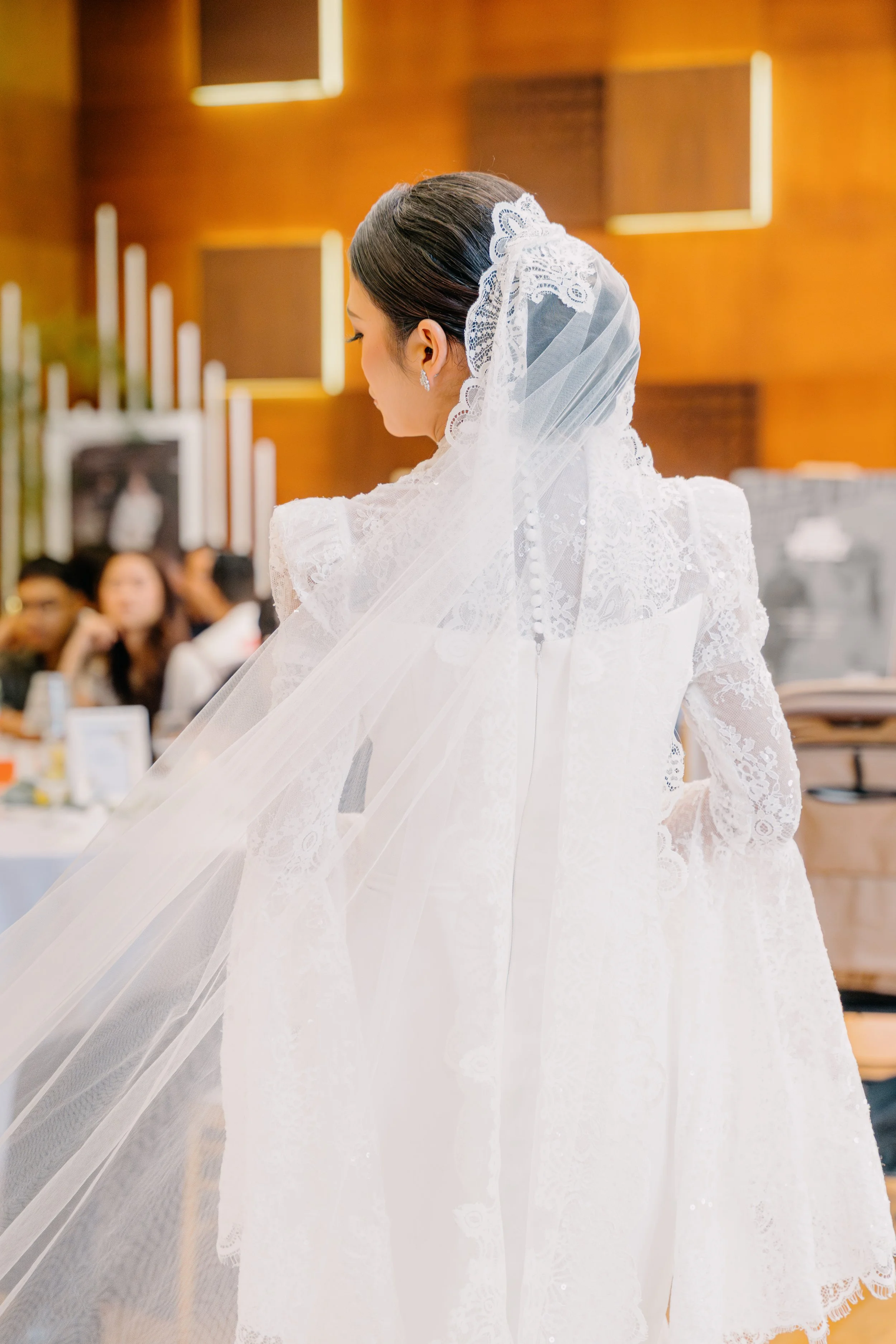 A bride in a white lace wedding dress with a lace veil standing in a warmly lit room with wooden panel walls, with guests seated in the background.