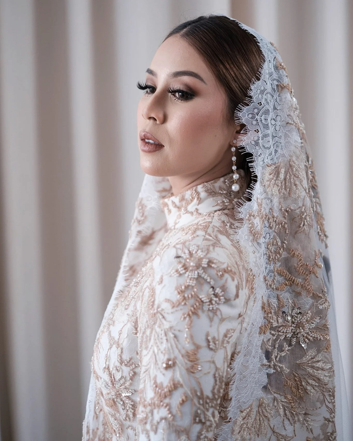 A woman in elegant wedding attire with detailed gold and white lace on her dress, wearing pearl earrings and a lace veil with floral accents, posing in front of a neutral backdrop.