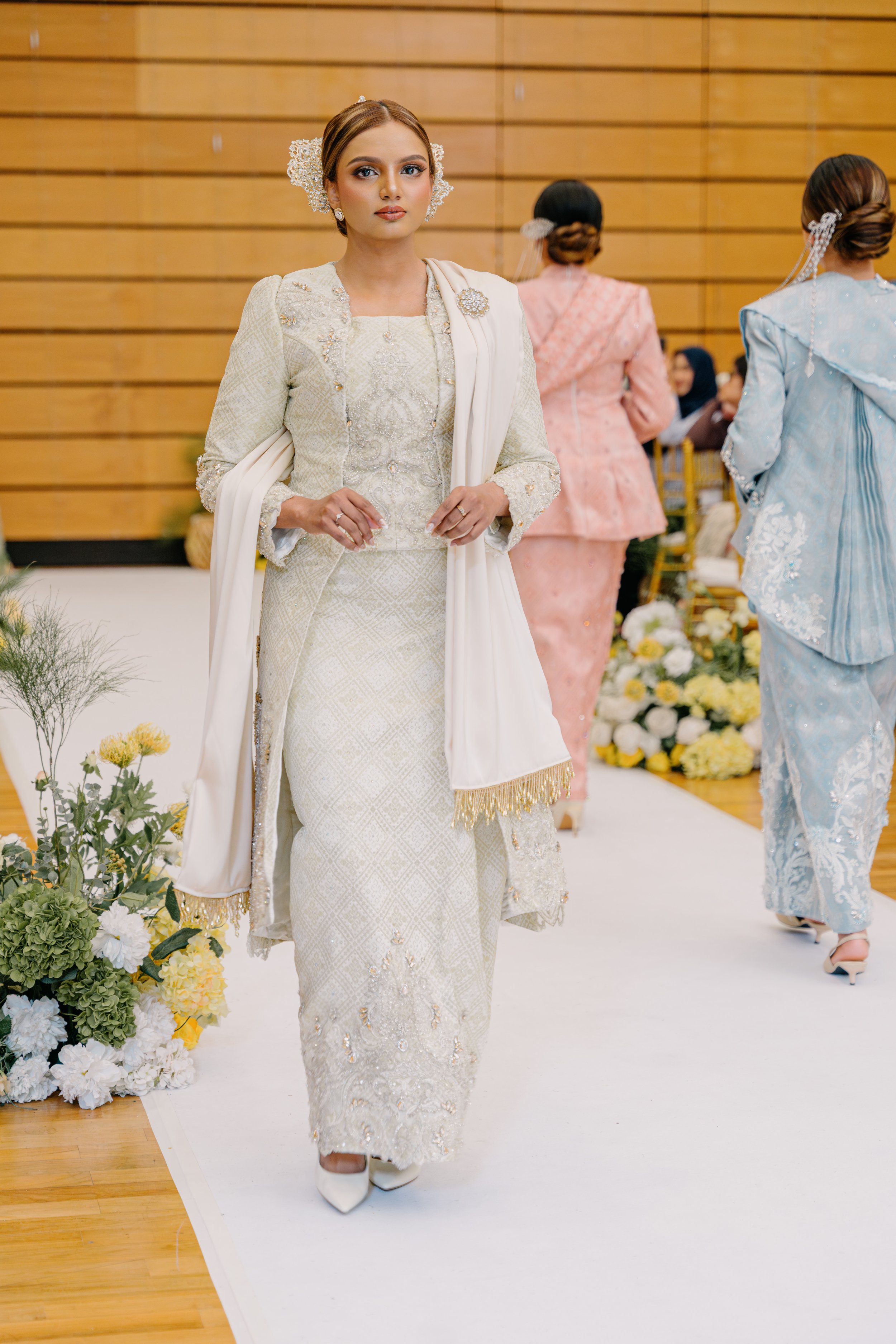 A woman in a traditional ornate white dress walking on a white carpet at a wedding ceremony, with floral arrangements on the side and women in colorful dresses in the background.