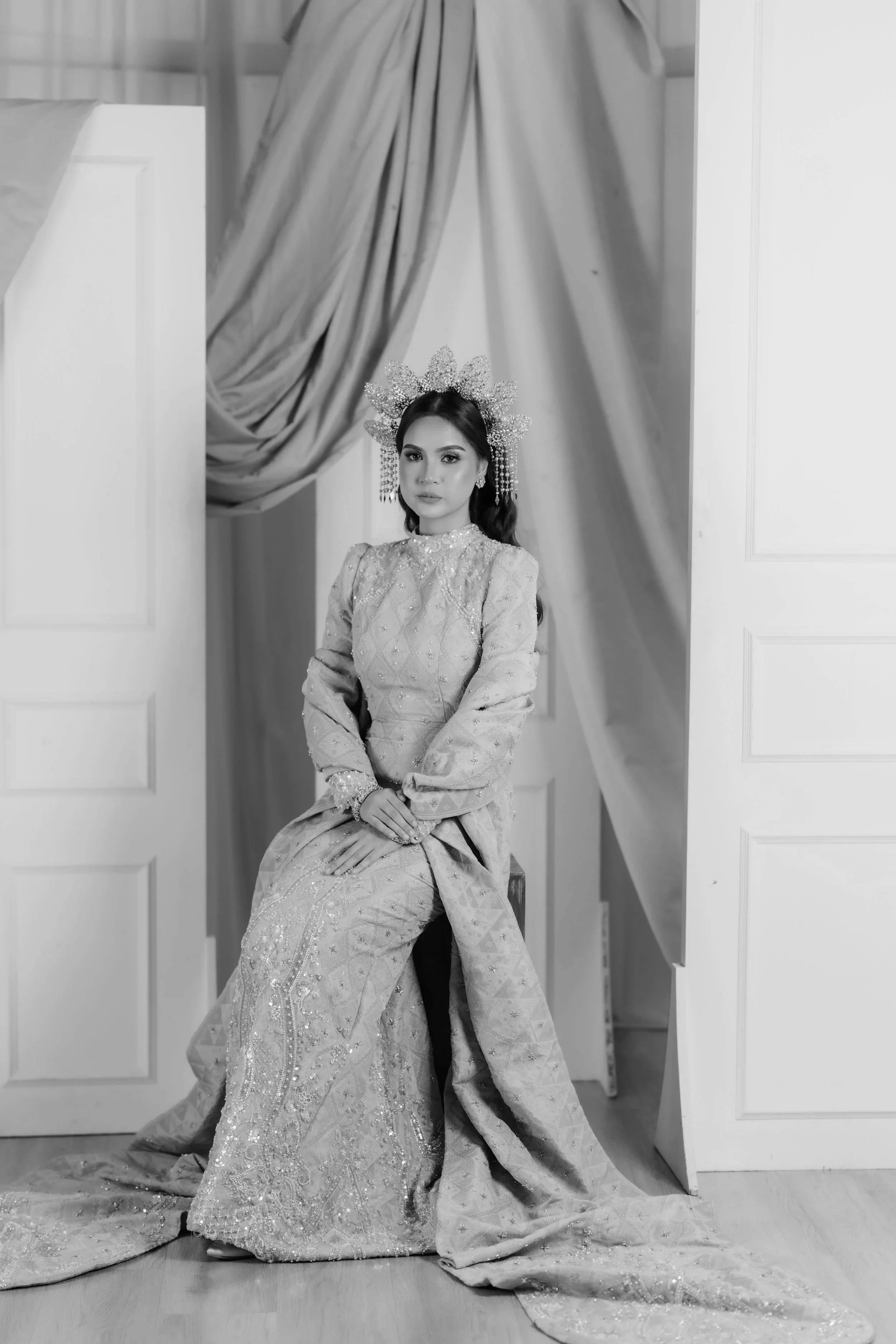 Black and white photo of a woman in a traditional, ornate gown and large, decorative headdress, seated with hands clasped, in a room with draped curtains and open double doors.