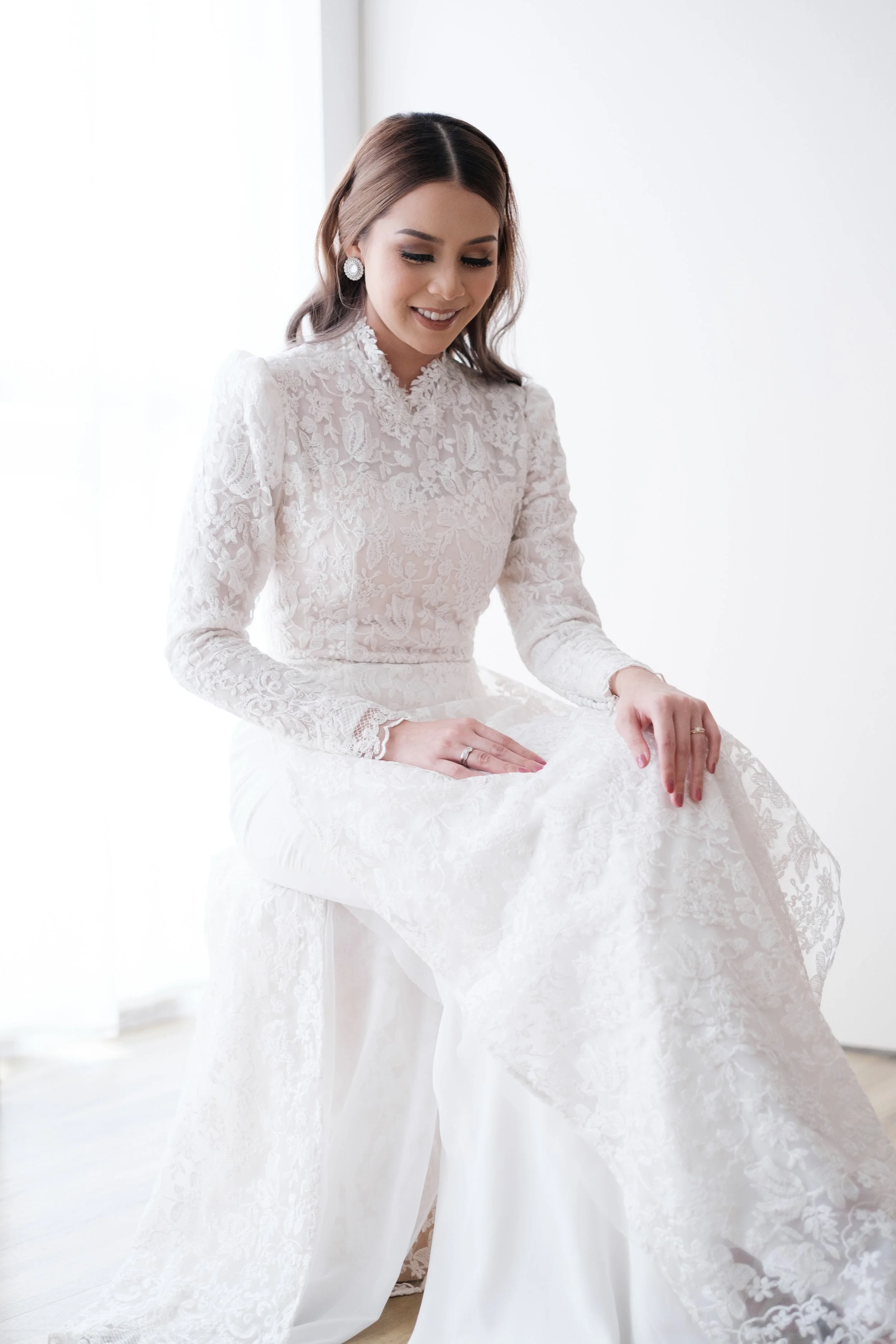 A young woman in a white lace wedding dress is sitting and smiling, with her eyes cast downward, in a bright, minimalist room.