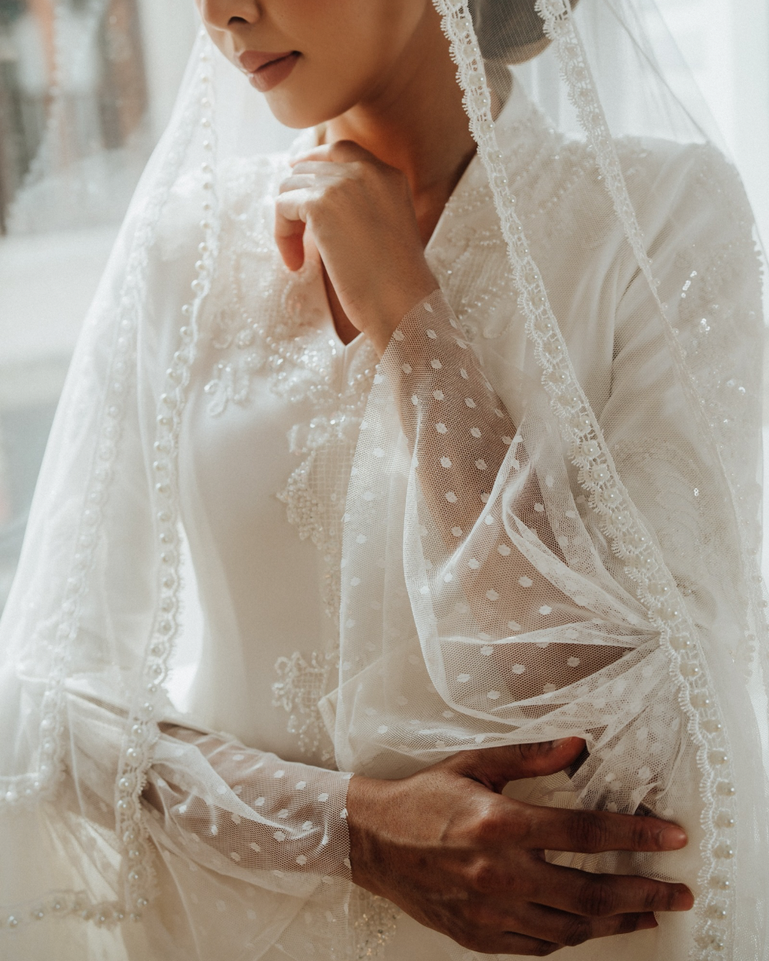 Close-up of a woman in a wedding dress with a lace veil, gently touching her chin and shoulder, with soft natural lighting.