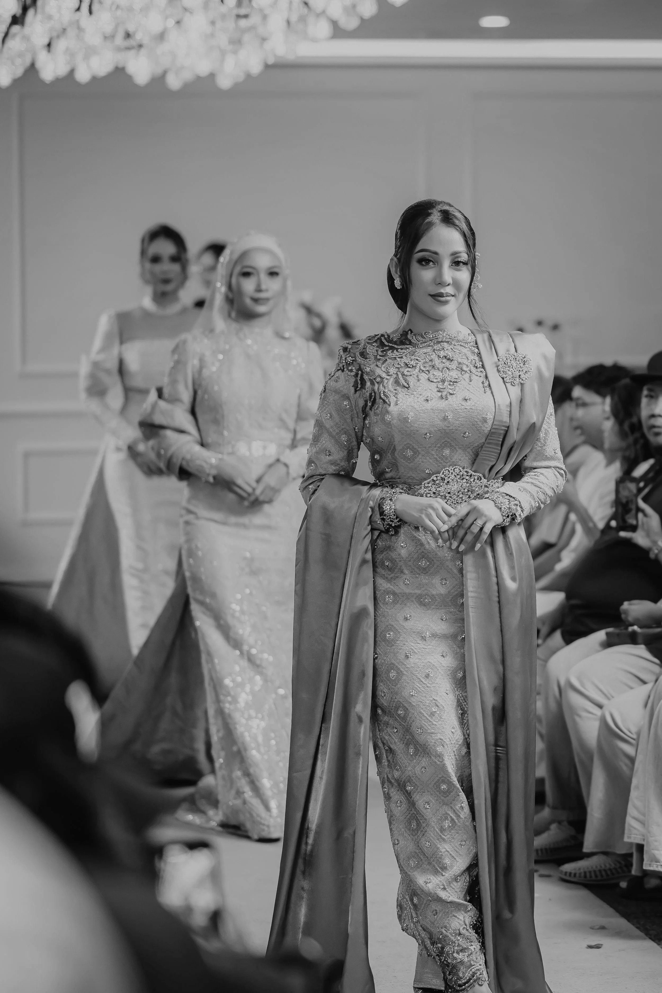 A woman in traditional attire walking down an aisle at a formal event, with seated guests in the background.