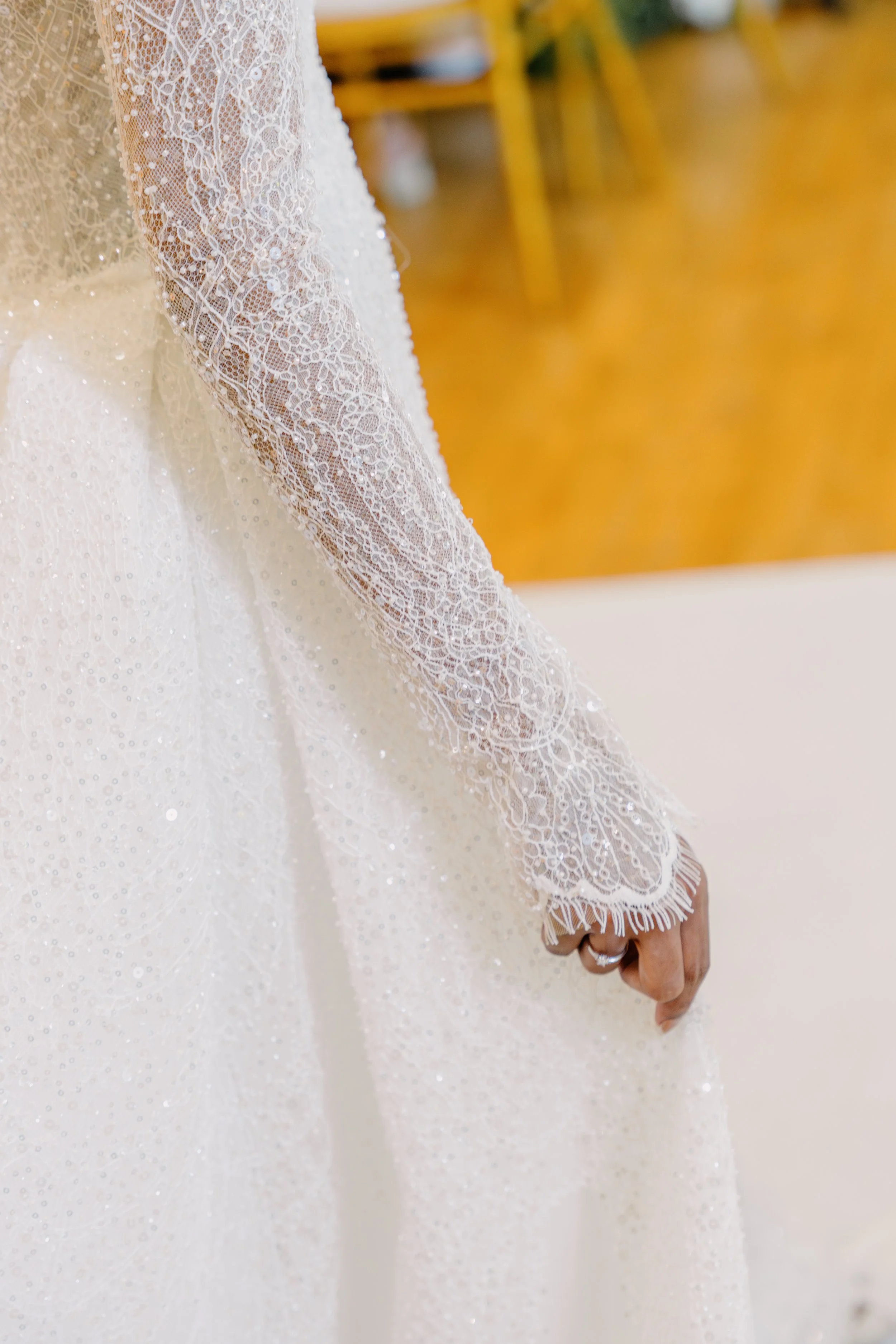 Close-up of a wedding dress with intricate lace and beading details, and a person wearing a ring on their hand.